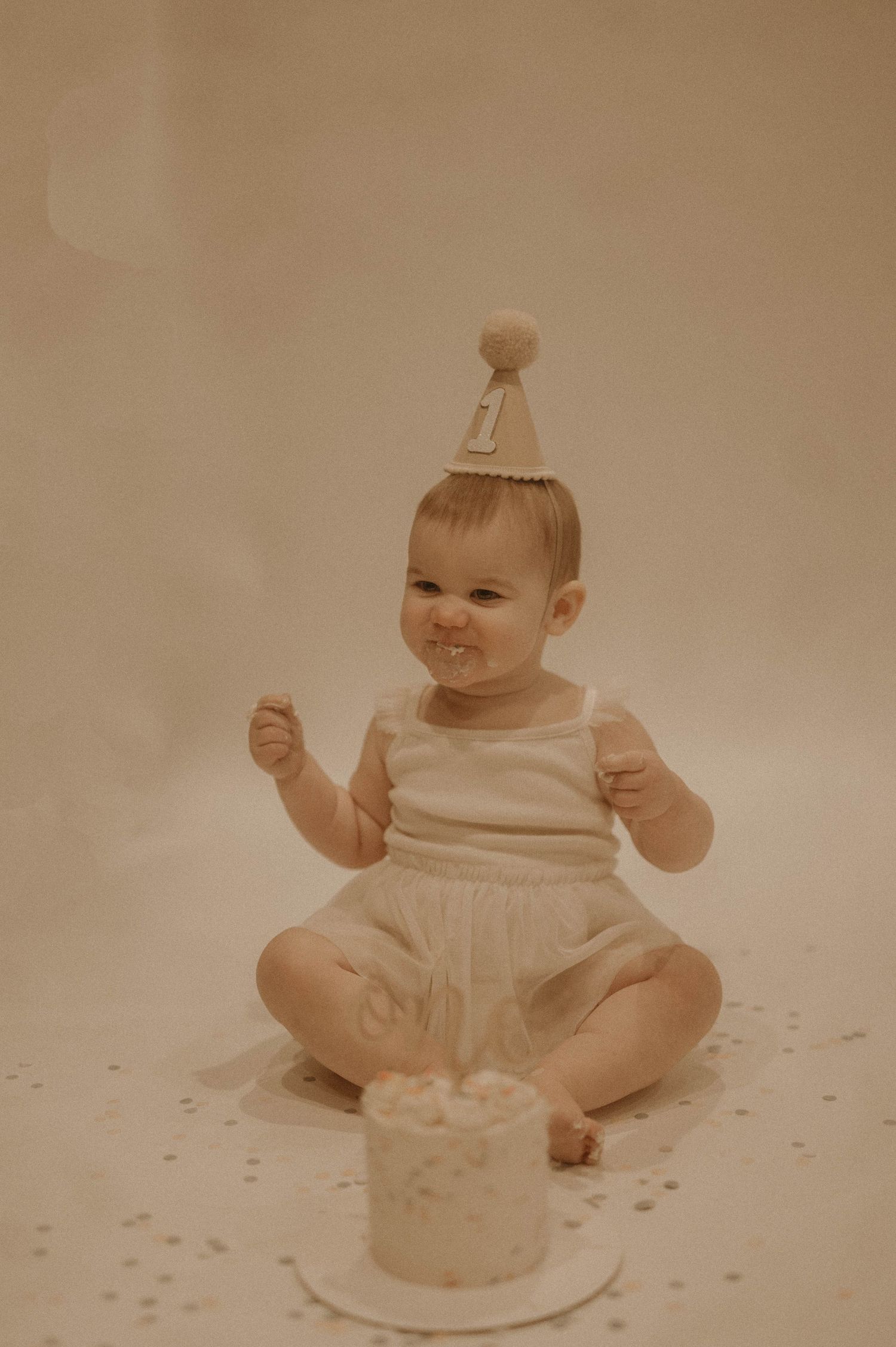 A baby in white clothing sits on the floor with a birthday cake, showing excitement and joy during their first birthday celebration.