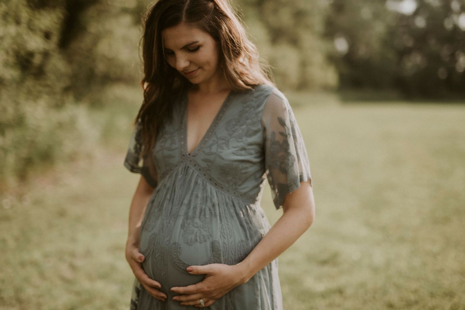 A person in a sage green dress cradles their pregnant belly in a sunny outdoor setting.