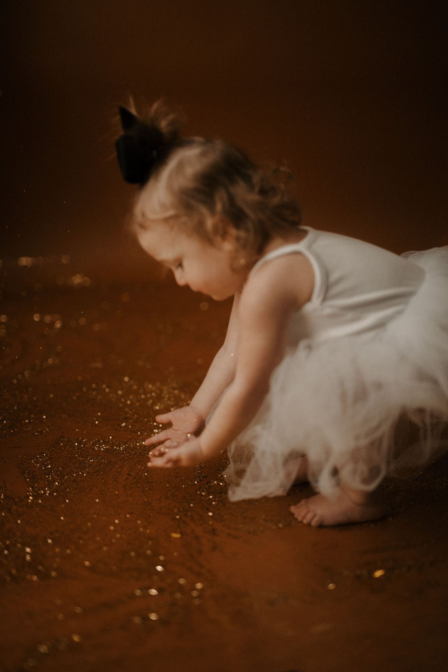 A small figure in a white tulle dress crawls on a dark floor with glittering sparkles scattered around.