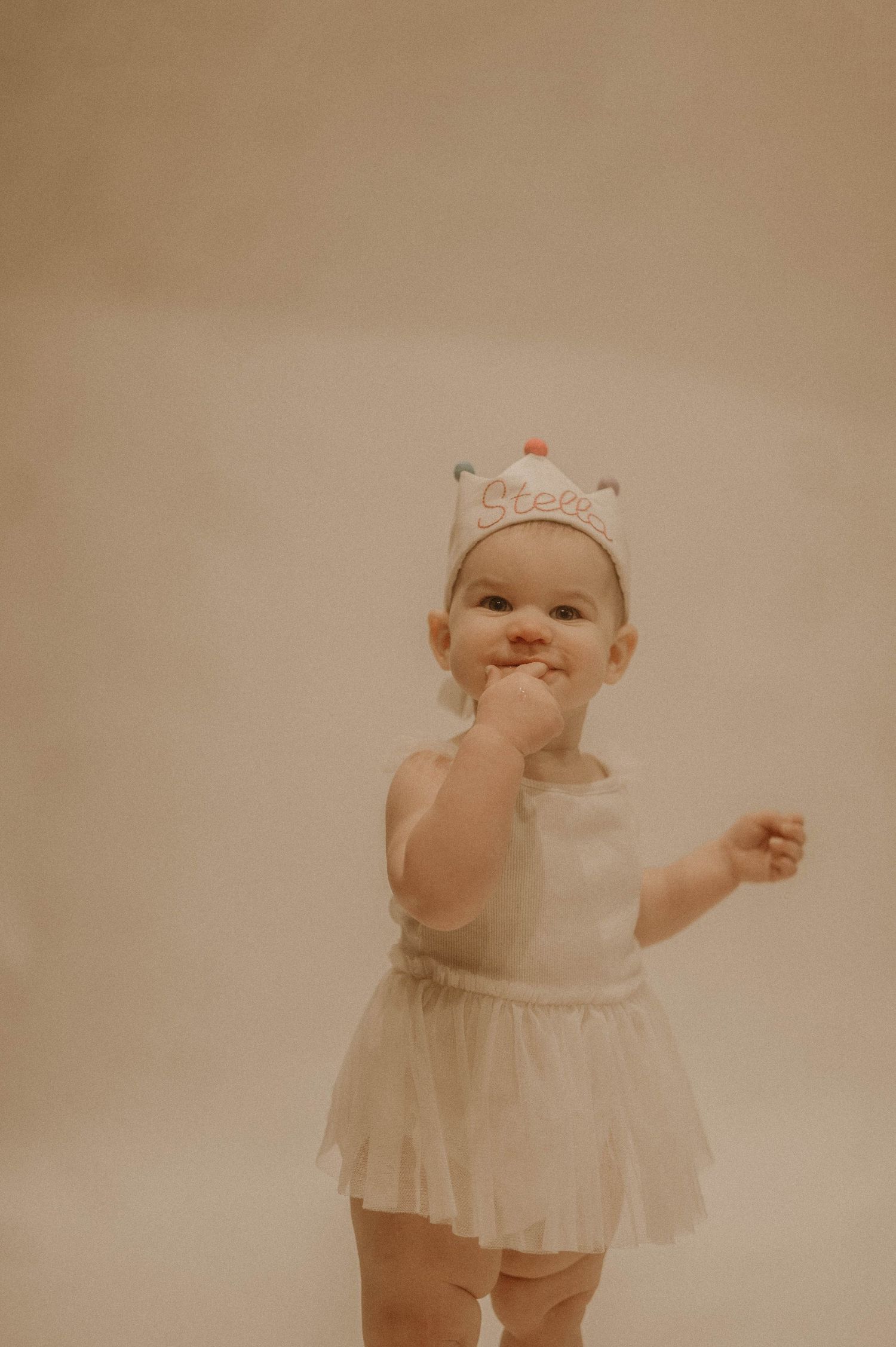 A small child wearing a white tutu dress and crown poses playfully against a neutral background.