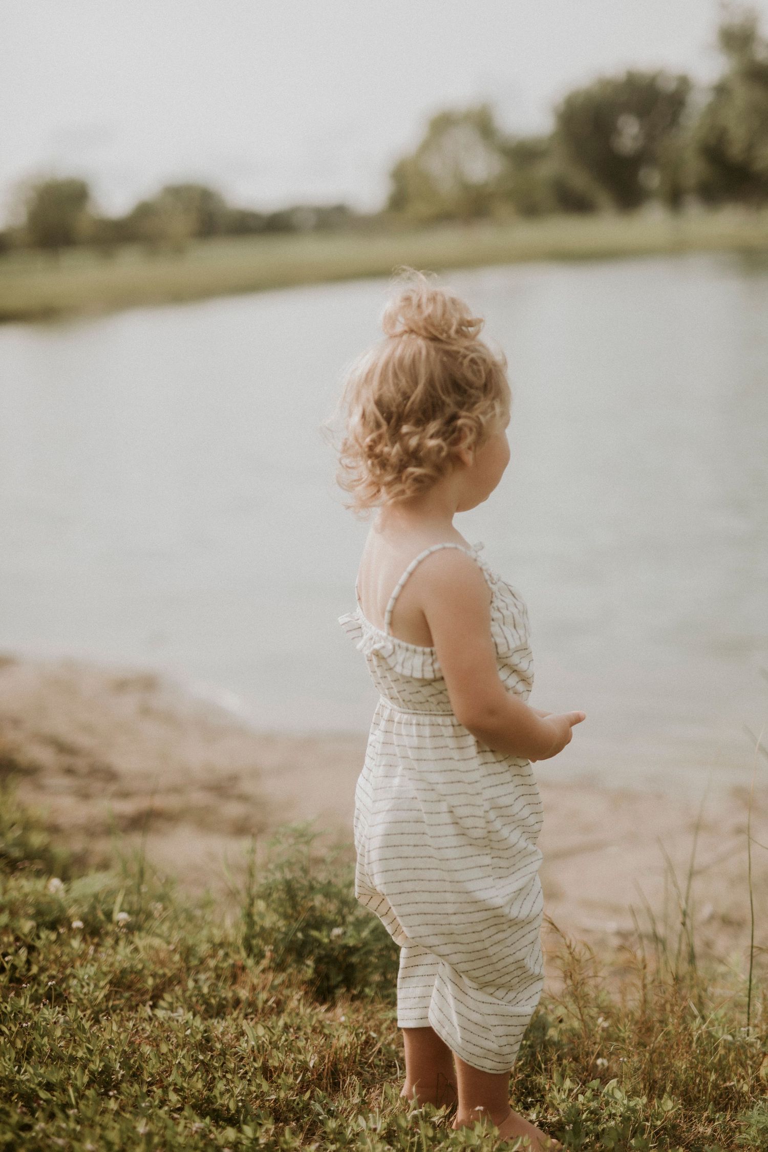 A small child in a striped white dress stands by a lake shore at sunset in a dreamy summer scene.