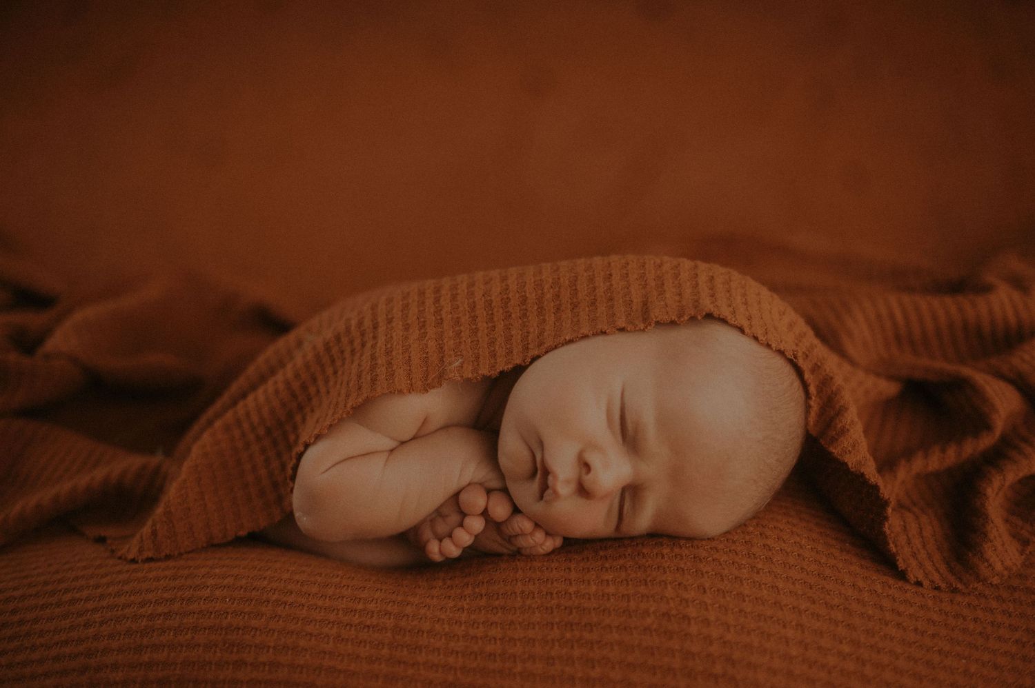 Sleeping newborn baby curled up peacefully under warm brown knitted blanket in soft lighting.
