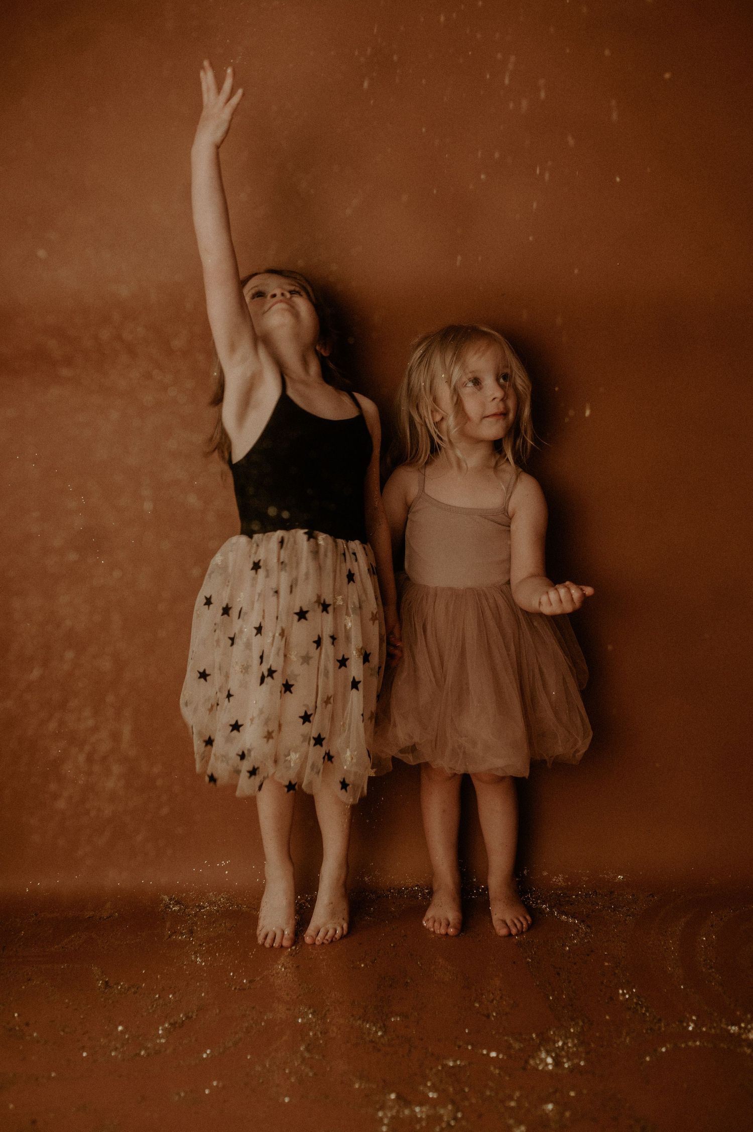 Two small children sit against a wall in sepia tones wearing polka dot and solid dresses.