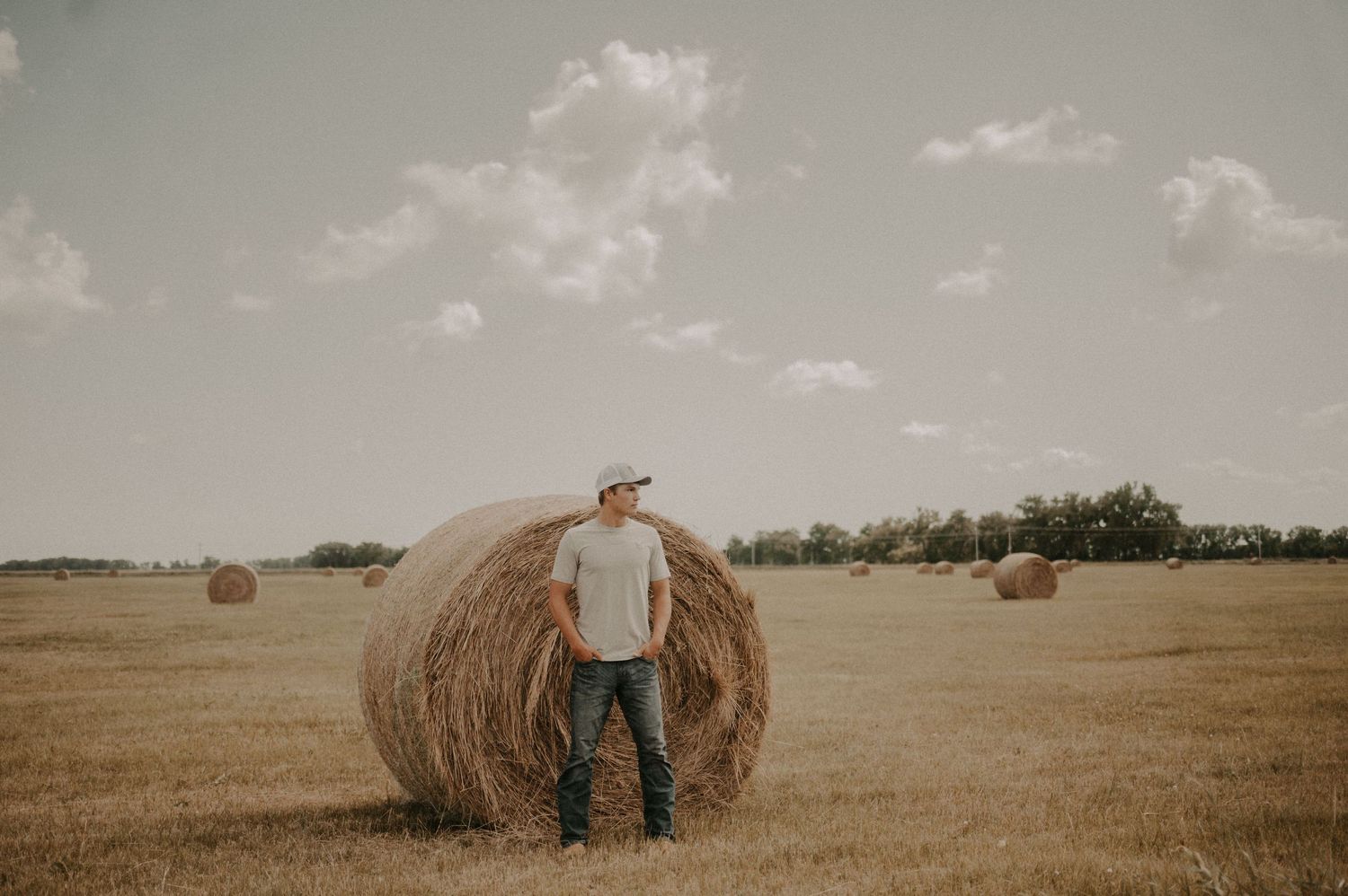 A man in a white t-shirt and cap stands beside a large hay bale in an open farm field under a partly cloudy sky.