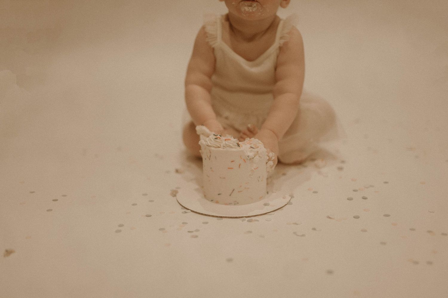 A baby wearing a white onesie sits on a floor with scattered crumbs or small objects around them.