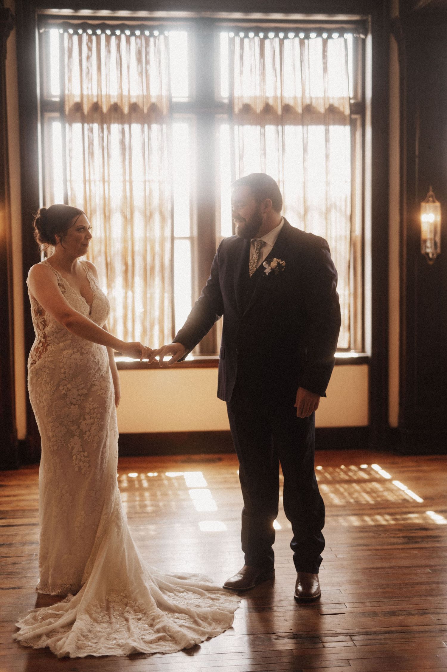 A wedding couple shares their first dance in a sunlit room with wooden floors and sheer curtains.