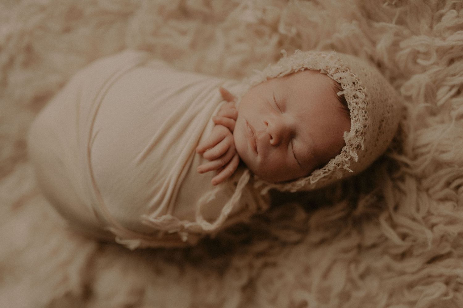 Sleeping newborn wearing lace bonnet resting on cream-colored fluffy rug.