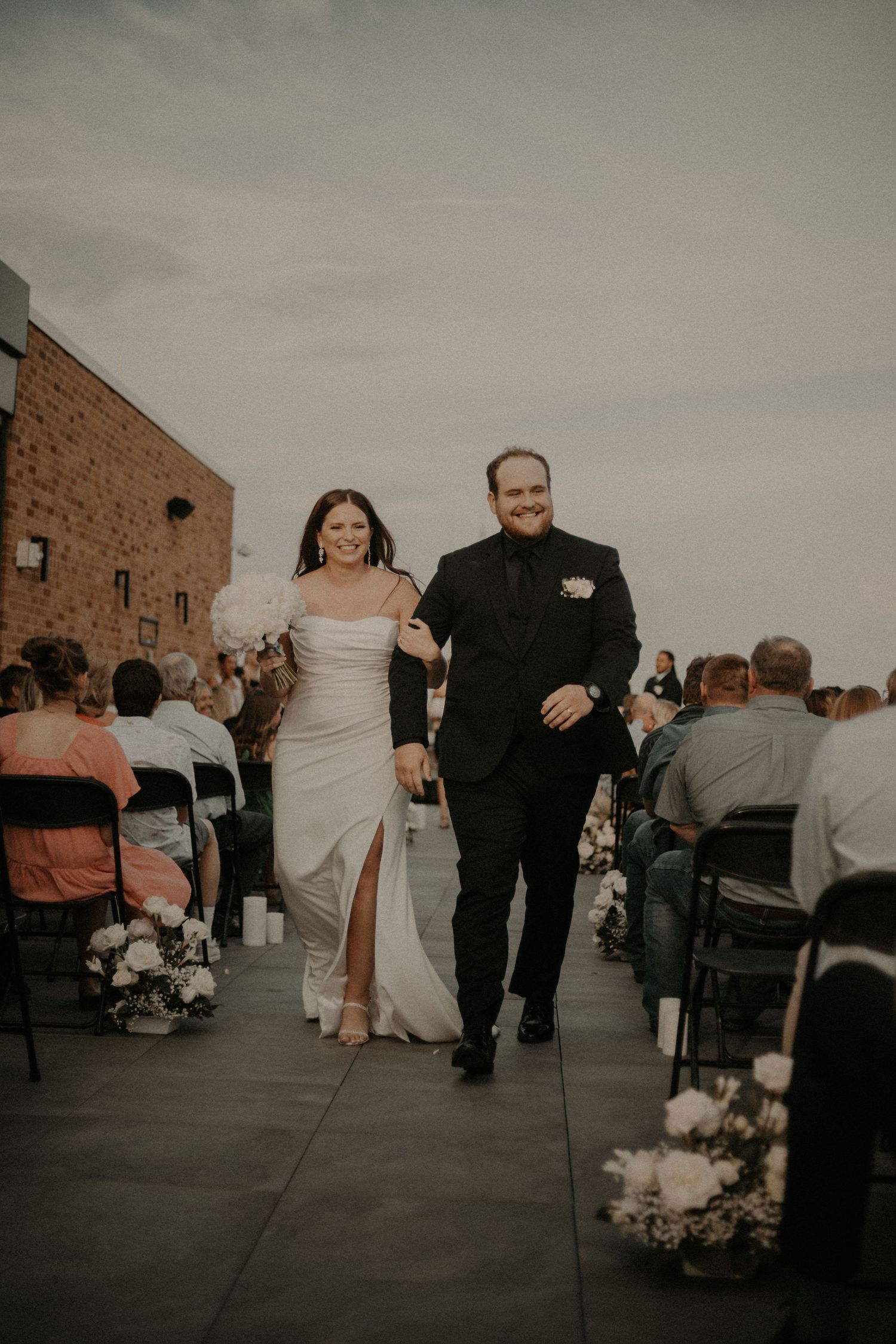 A bride and groom walk along a brick-lined path with wedding guests seated outdoors.