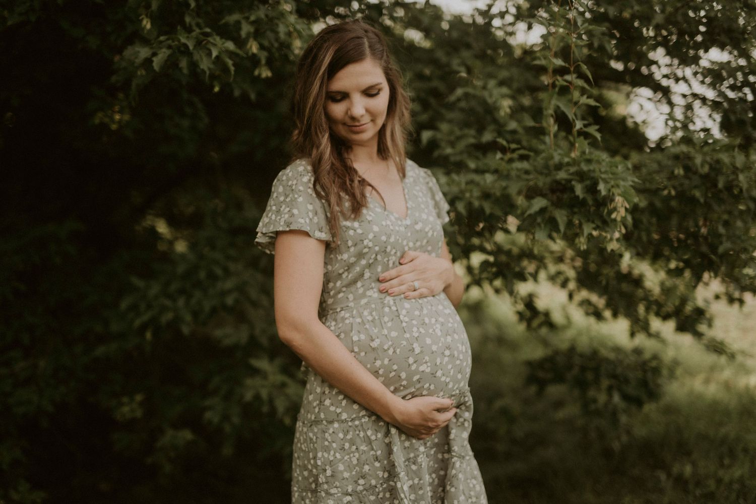 A person in a floral dress stands in a sunlit garden setting, cradling their pregnant belly with gentle affection.
