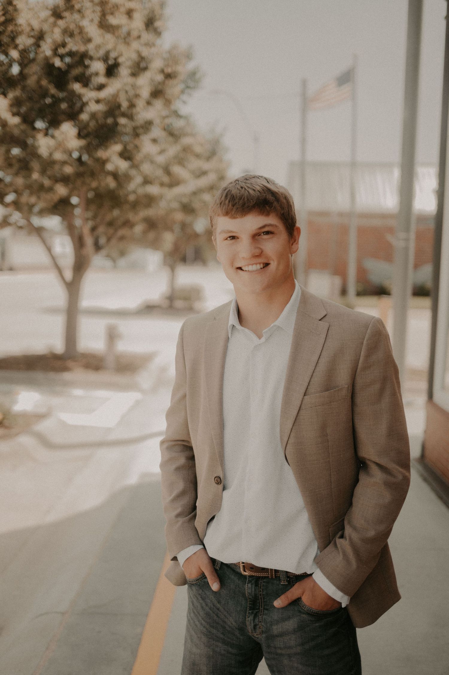 Professional portrait of someone in a tan blazer and white shirt standing outdoors with an American flag in the background.