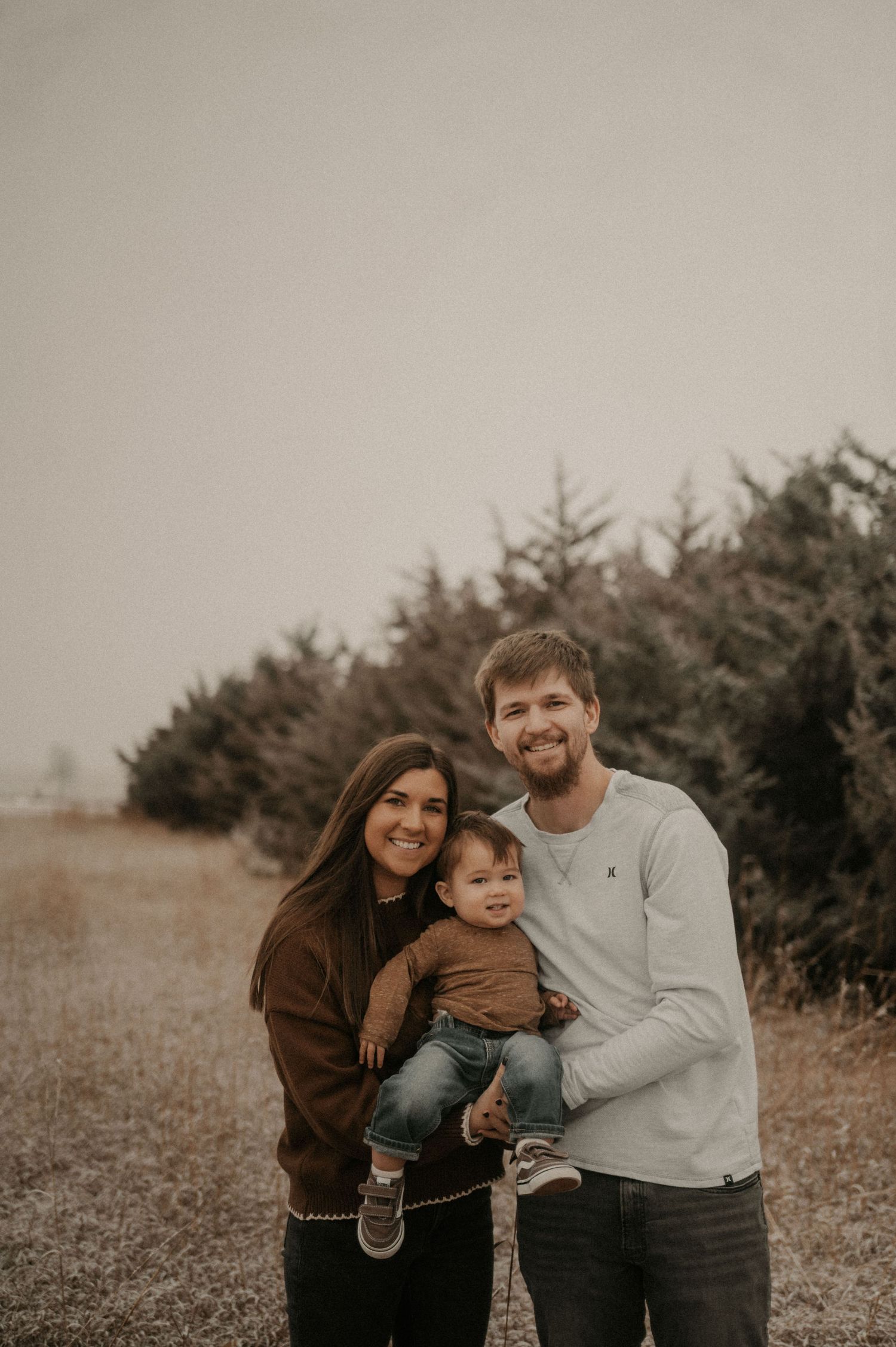 A family poses together outdoors in front of evergreen trees on an overcast day in warm earth-toned clothing.