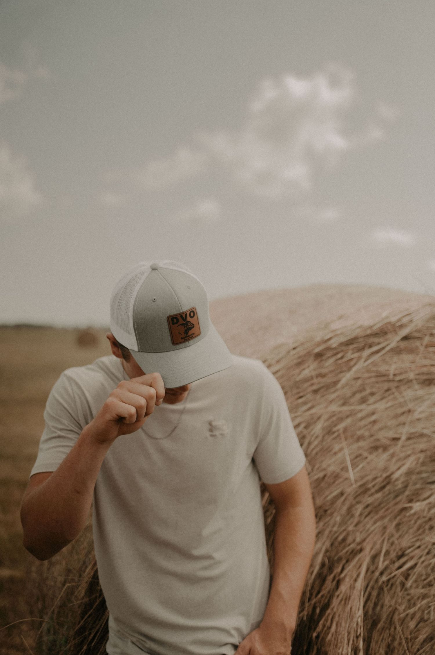 A person in a white t-shirt and baseball cap poses against a golden hay bale in moody outdoor lighting.