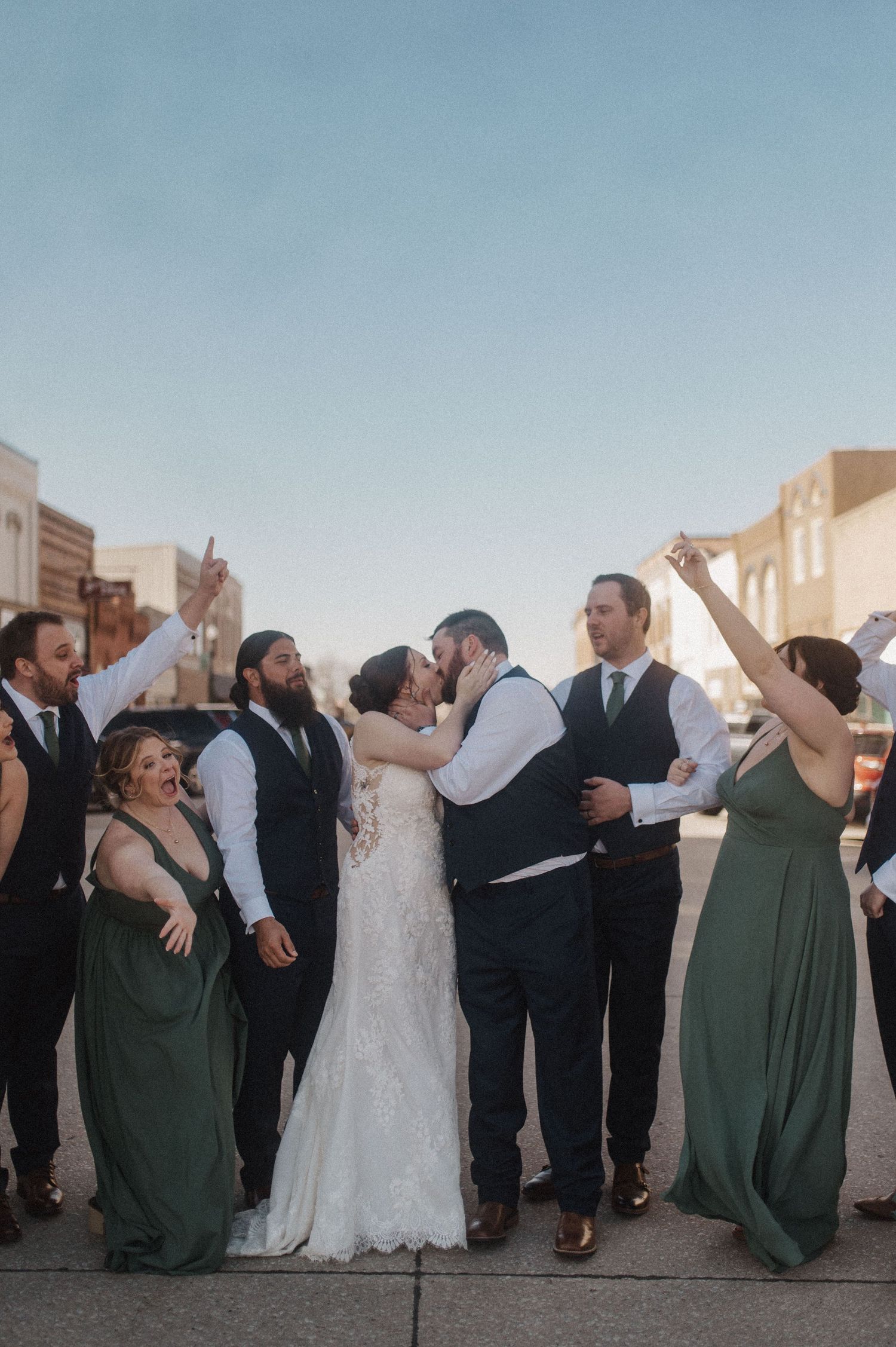 Wedding party in green dresses and suits posing outdoors at sunset.