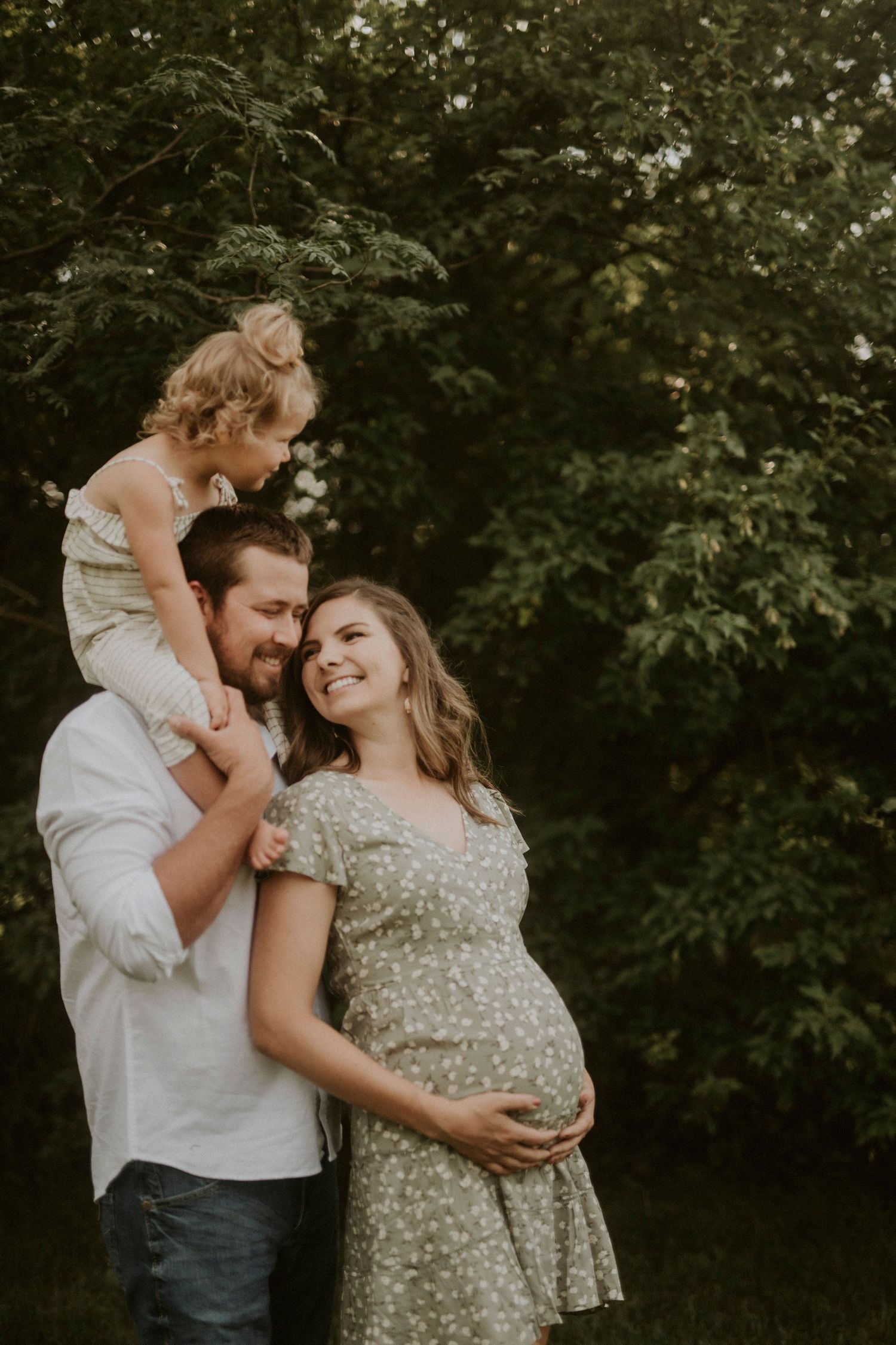 A couple shares intimate moments in an outdoor maternity photoshoot against a backdrop of lush greenery.
