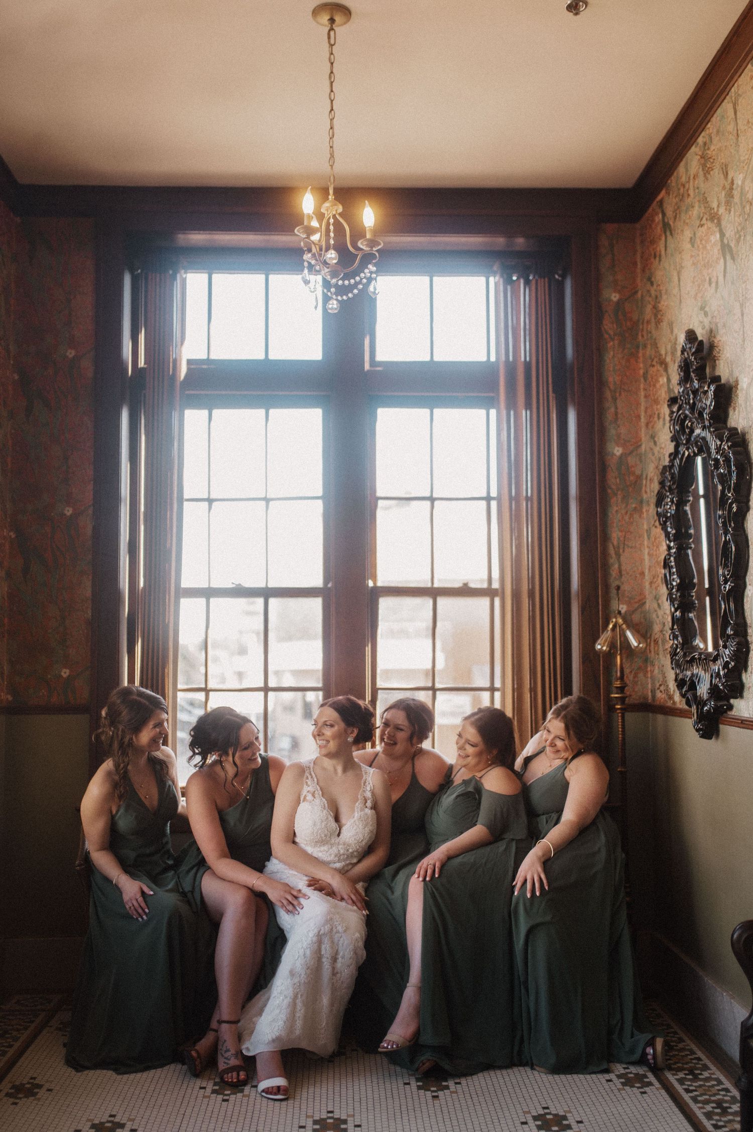 Bridesmaids in emerald green dresses sit together by large windows in an elegant room.