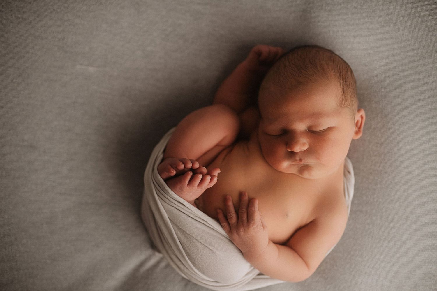 Newborn baby sleeping peacefully while wrapped in light fabric on gray backdrop.