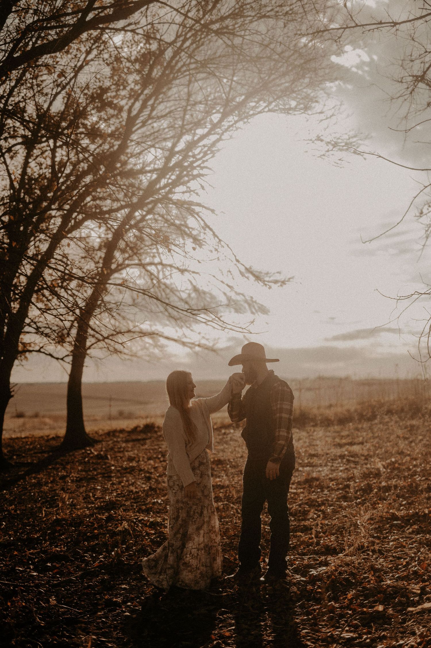 Two people sharing an intimate moment among fallen leaves and trees at dusk.