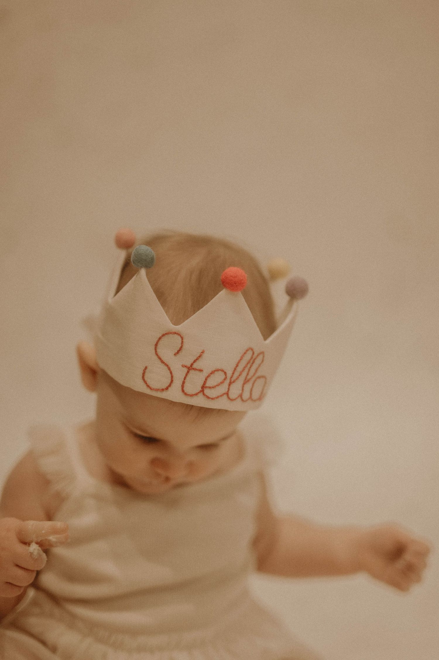 A baby wearing a white cloth crown lies on a beige background in a soft, dreamy portrait series.
