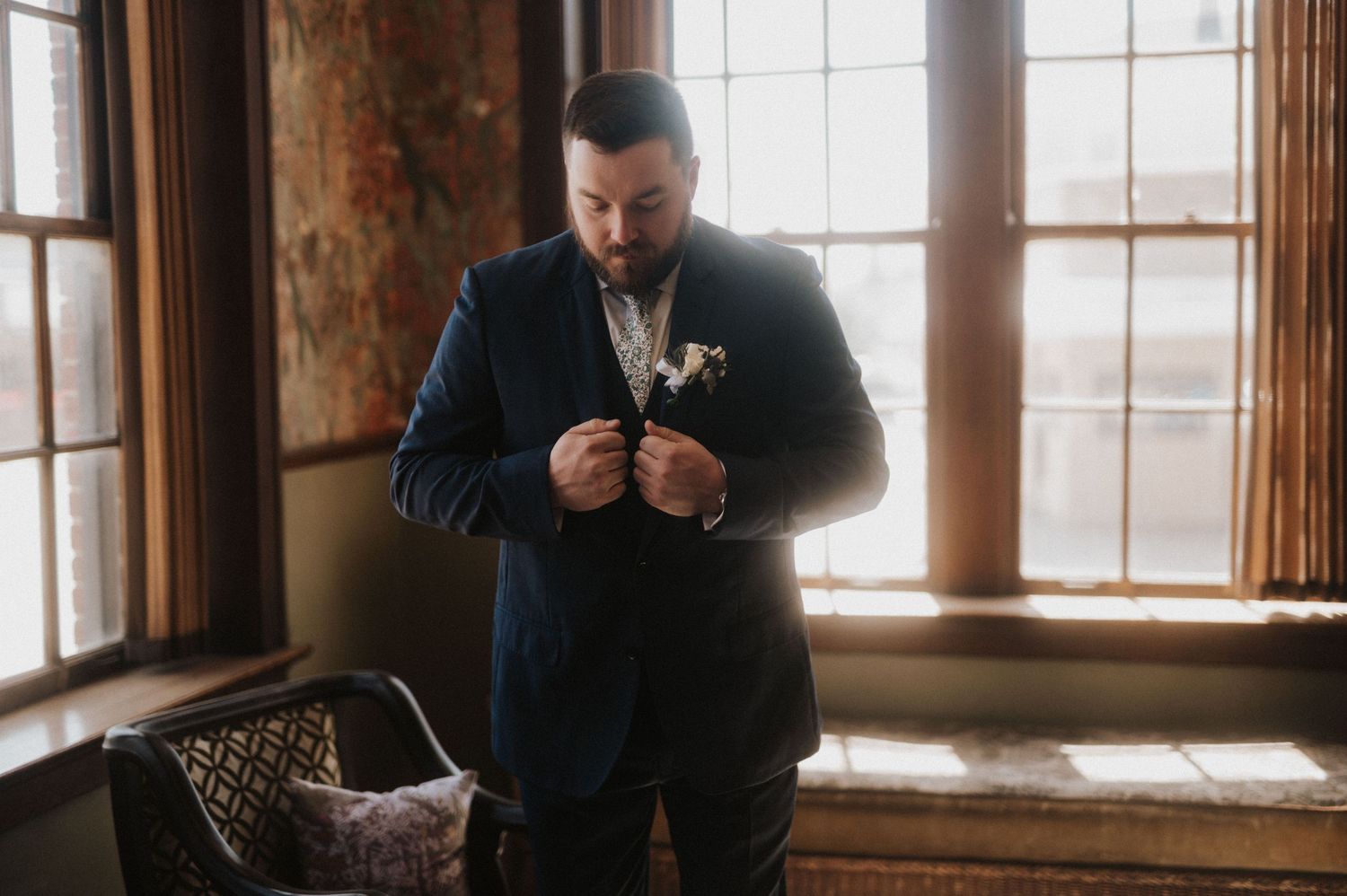 A person in a dark suit adjusts their tie while standing near large windows in a rustic room with natural light.