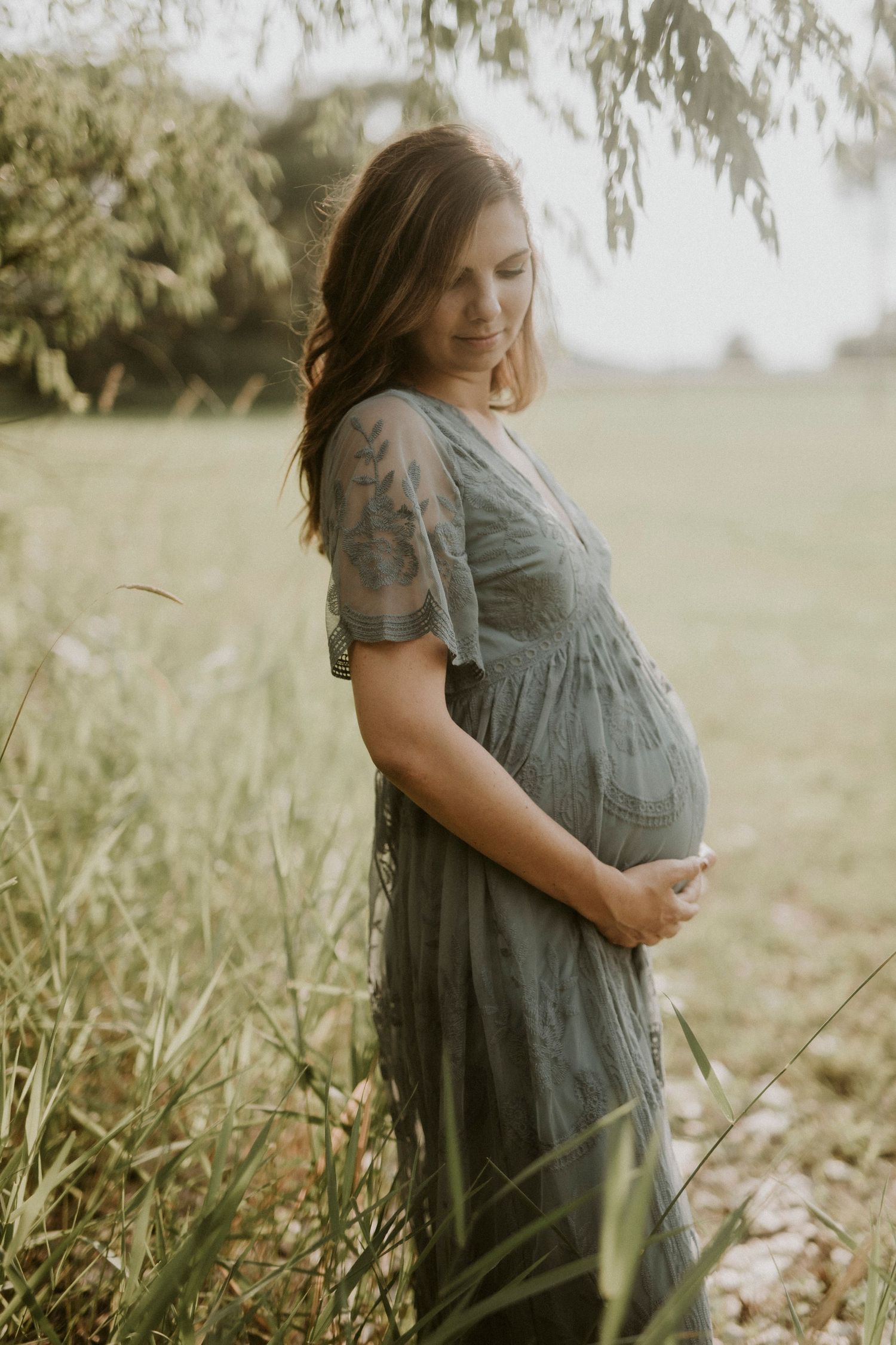 A pregnant woman in a grey dress stands thoughtfully in a sunlit meadow with willow branches overhead.
