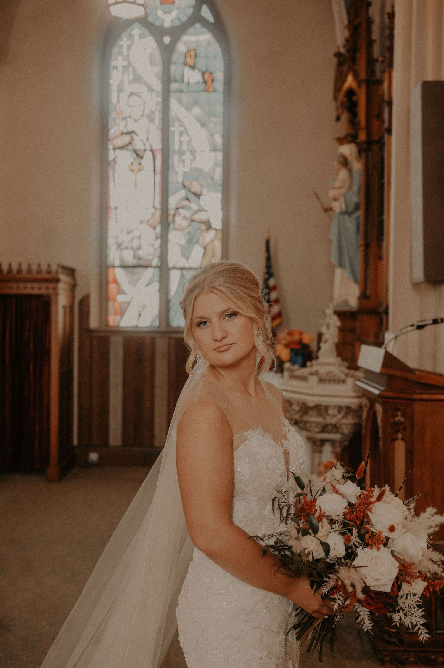 Bride holds white and burgundy floral bouquet in front of stained glass church windows.