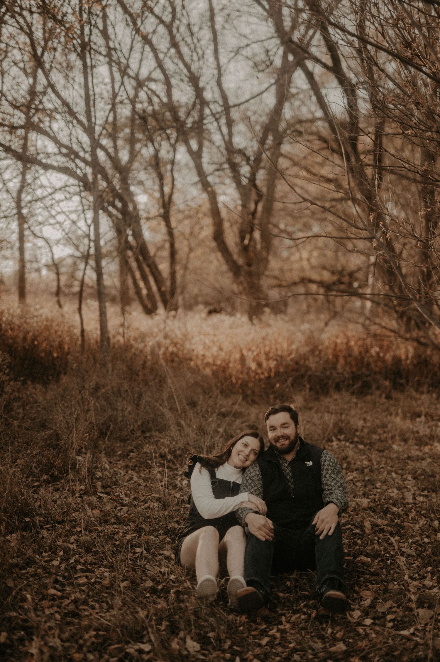 A couple sits together on fallen autumn leaves in a moody forest setting during golden hour.