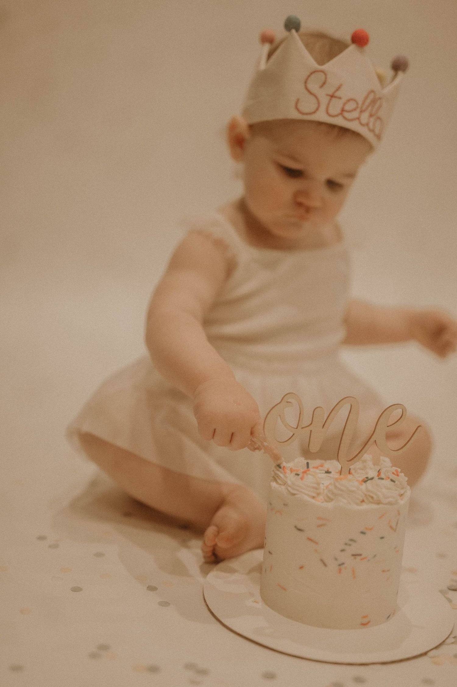 Baby in white reaching for a small pink birthday cake with sprinkles on a light beige background.