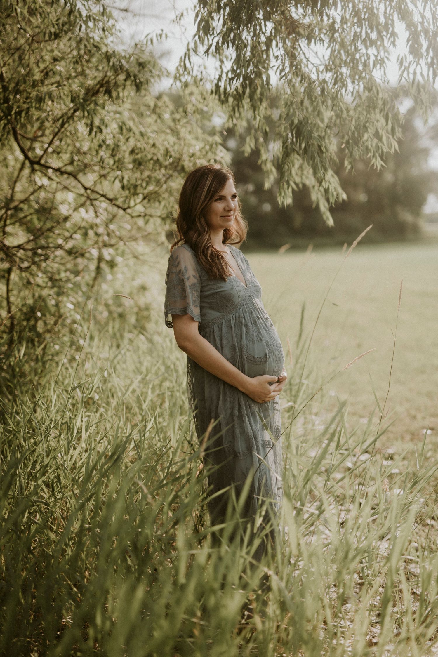 A person in a flowy dress stands among tall grass and weeping willow trees during golden hour.