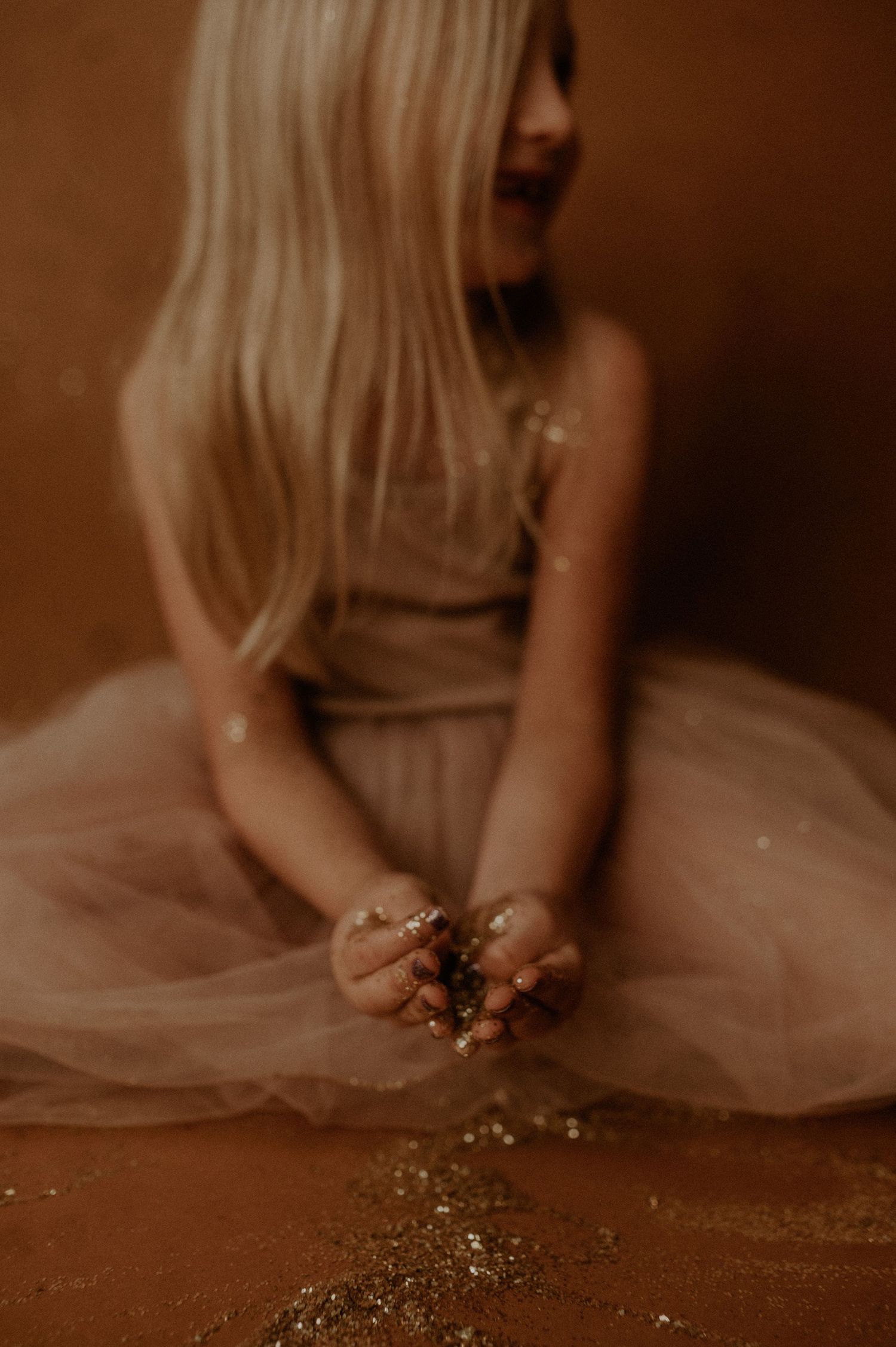 A person in a beige dress sits on a wooden floor while gold glitter sparkles and scatters around them.