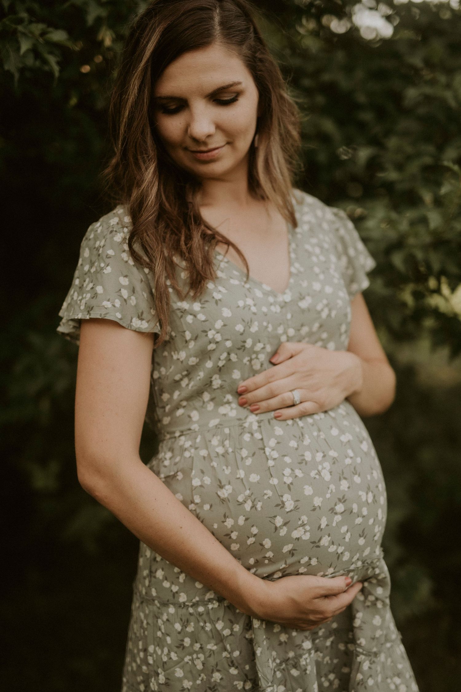 A pregnant woman in a floral dress tenderly holds her baby bump while standing in a garden setting during golden hour.