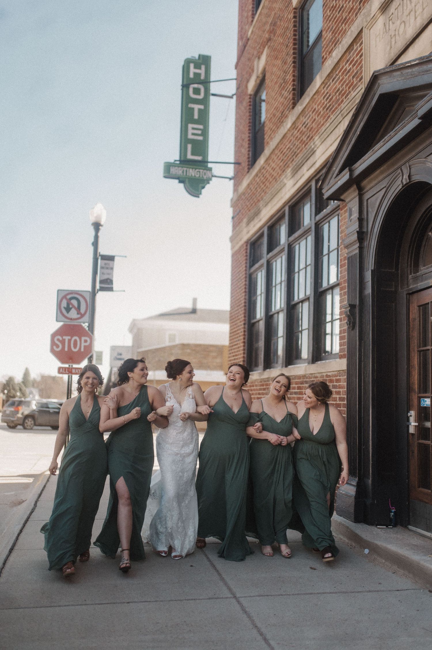 Bridal party in green dresses sitting along sidewalk near hotel sign.