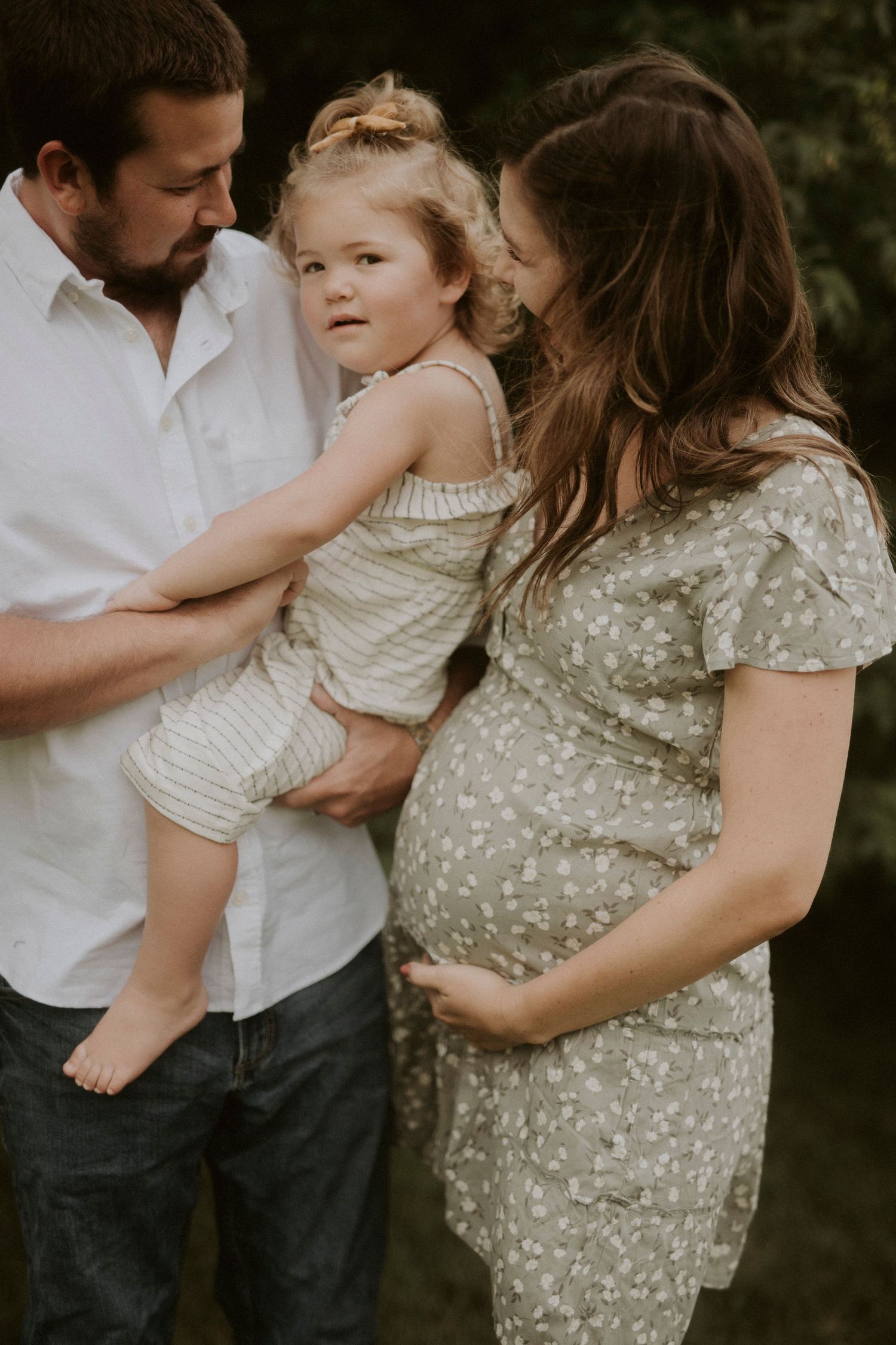 Intimate family maternity photo outdoors showing couple in white and floral dress holding small child in cream romper.