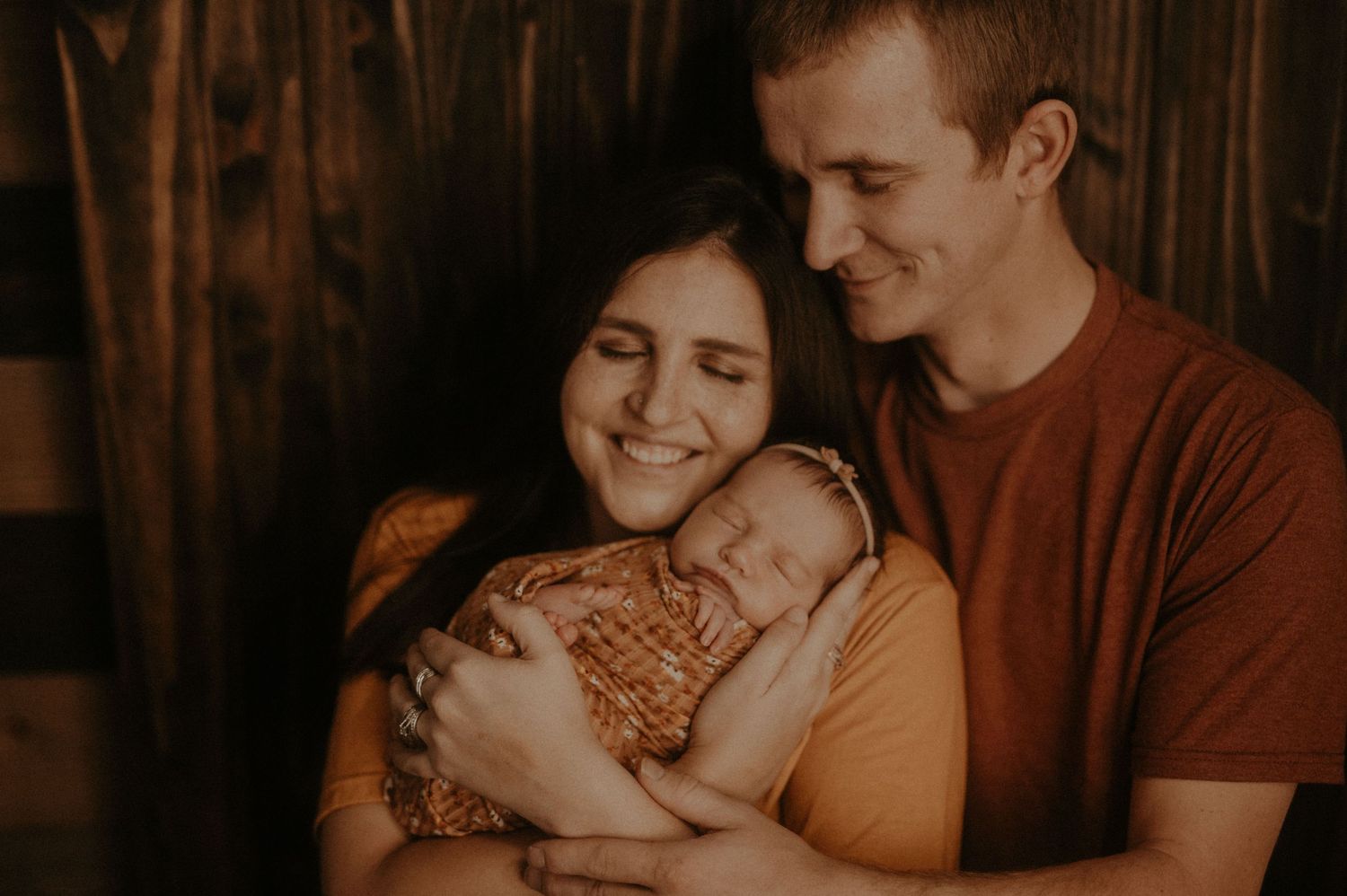 A couple smiles warmly while holding their sleeping newborn baby in a cozy indoor setting with warm lighting.