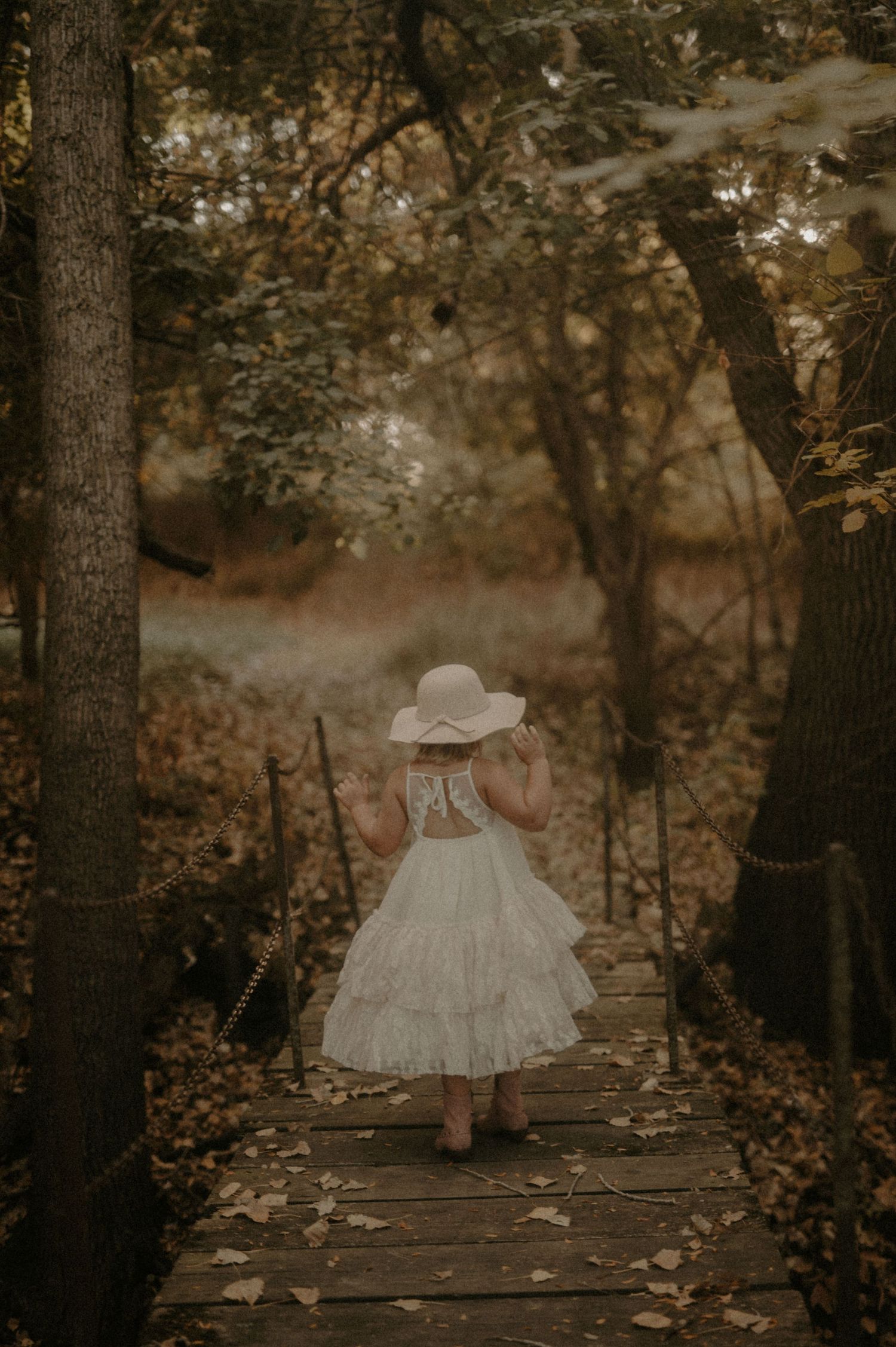 Small child in a white dress walking along a wooden path through an autumn forest.