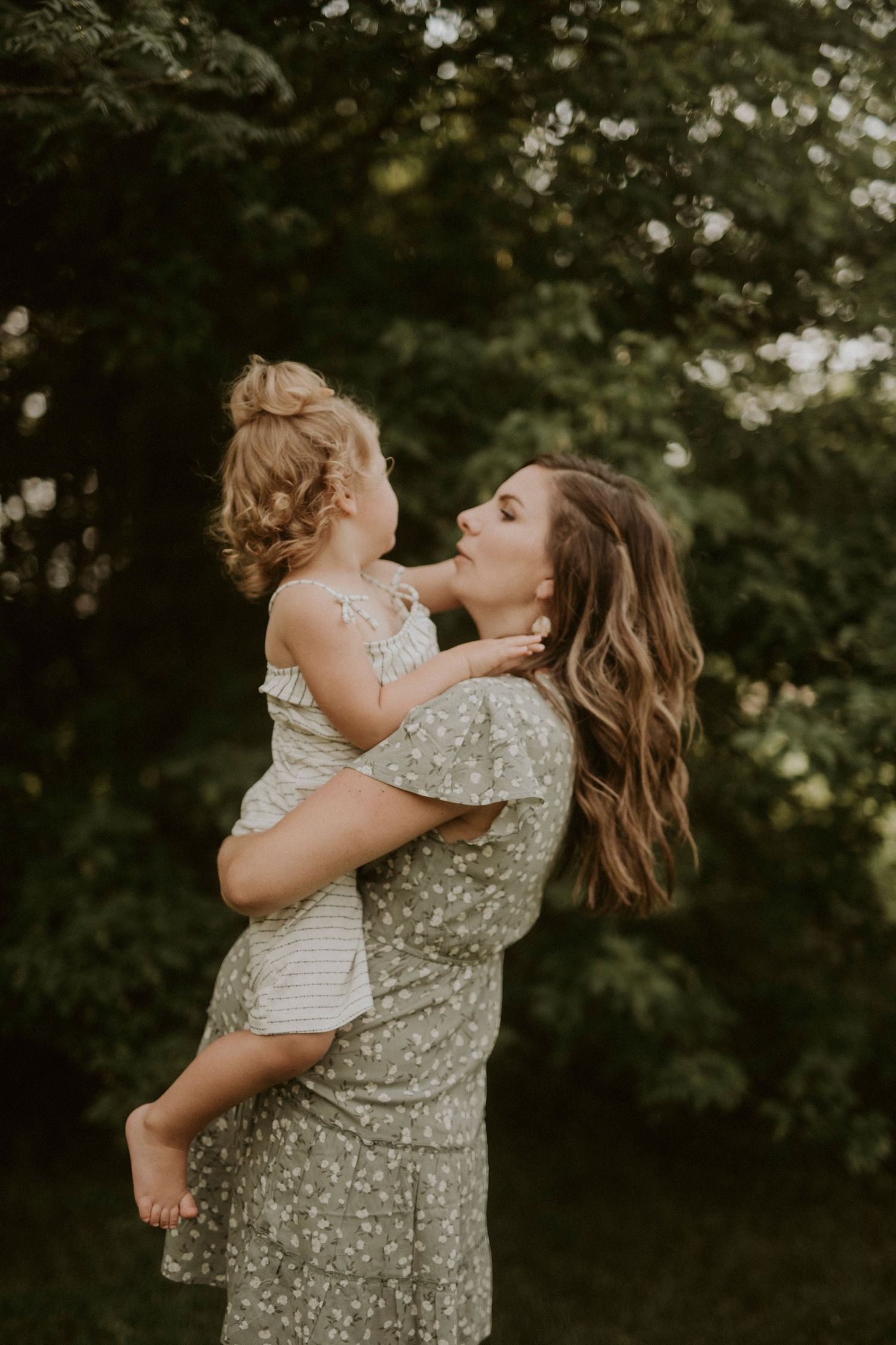A tender moment captured outdoors as a parent in a floral dress lifts and embraces a small child against a green forest backdrop.