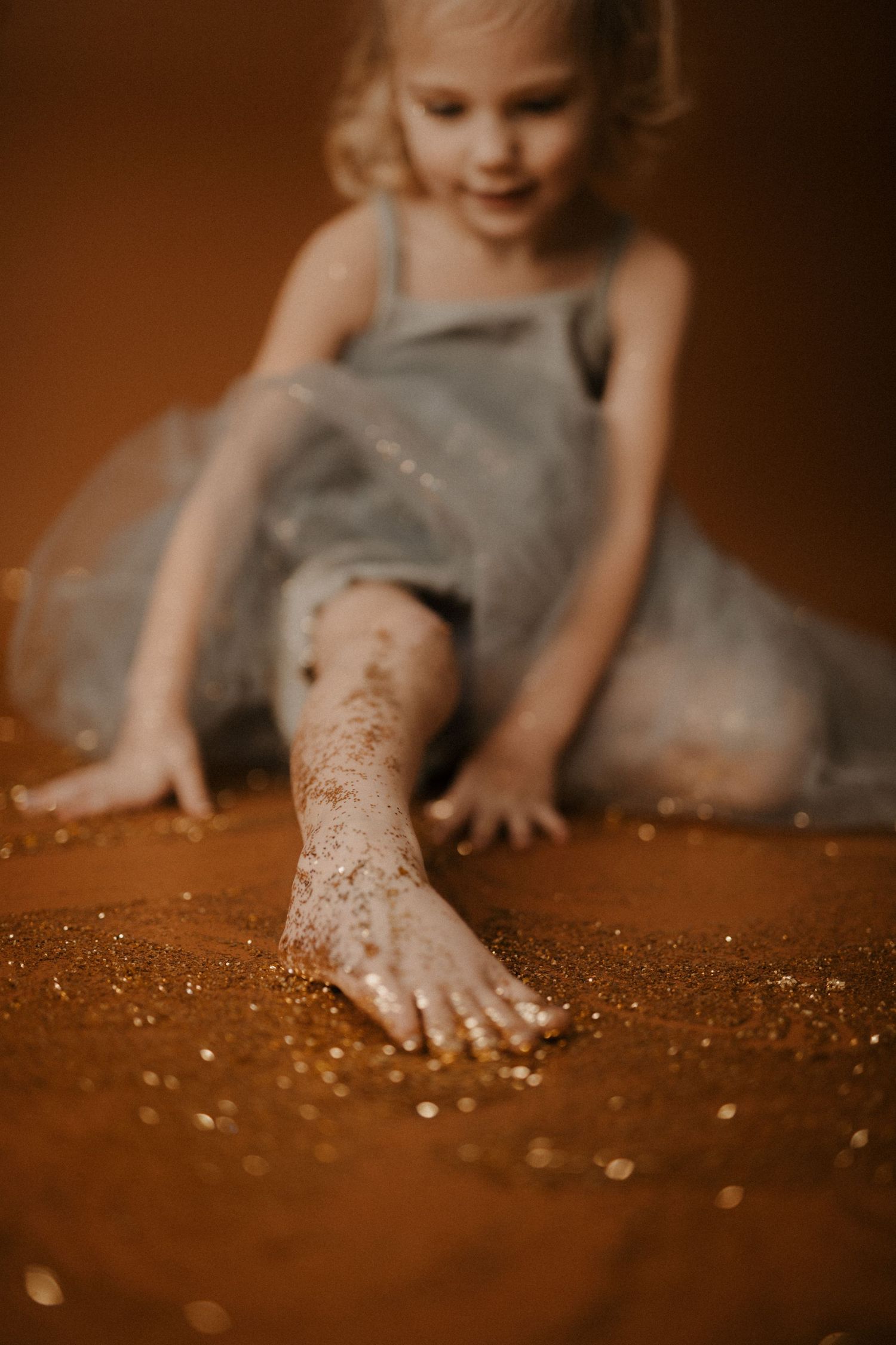 Child in gray tulle dress plays with glitter on wooden floor creating magical sparkly moment.