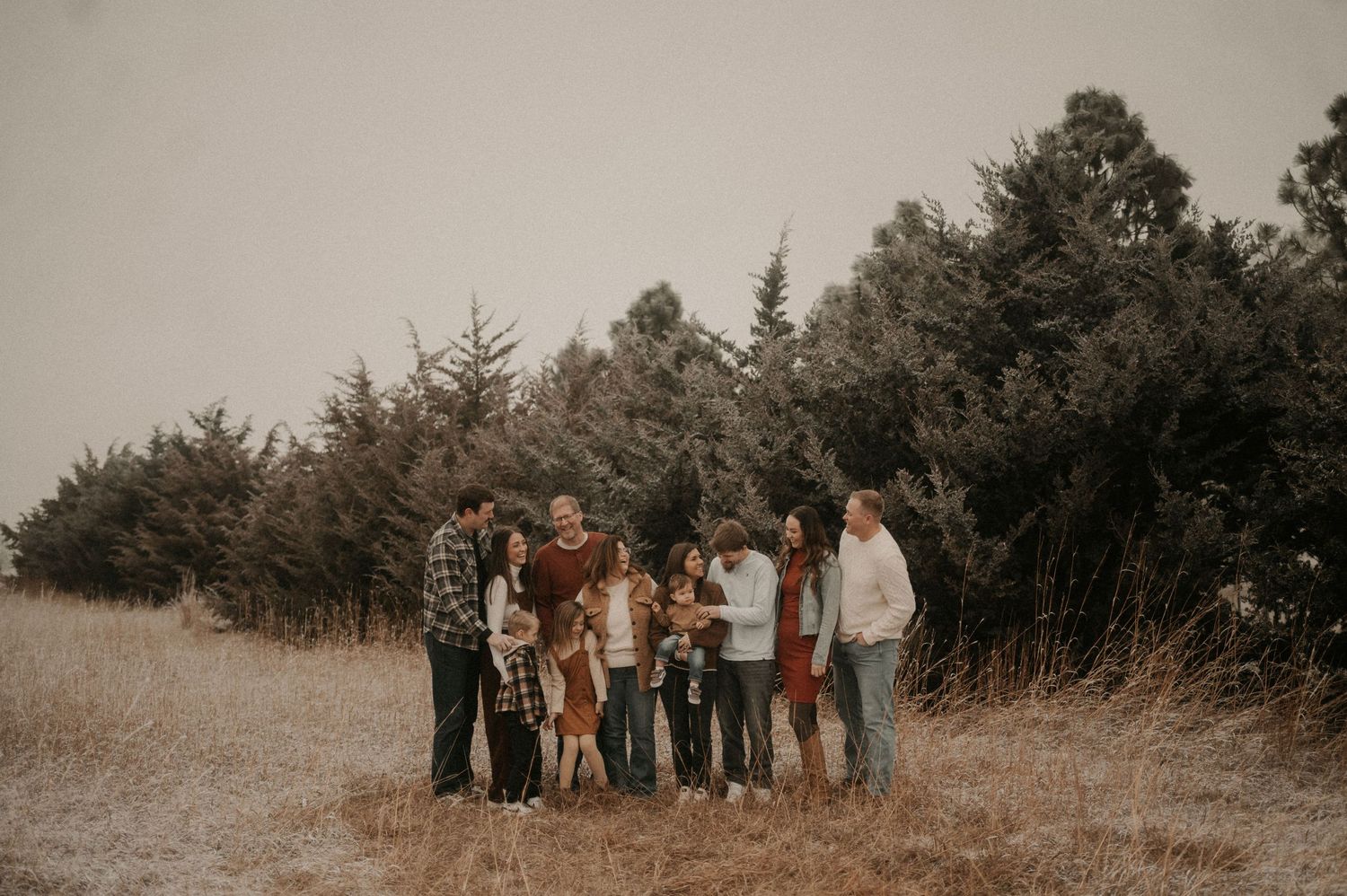 A group of people stand together in a line against a rustic outdoor backdrop with trees and natural scenery.