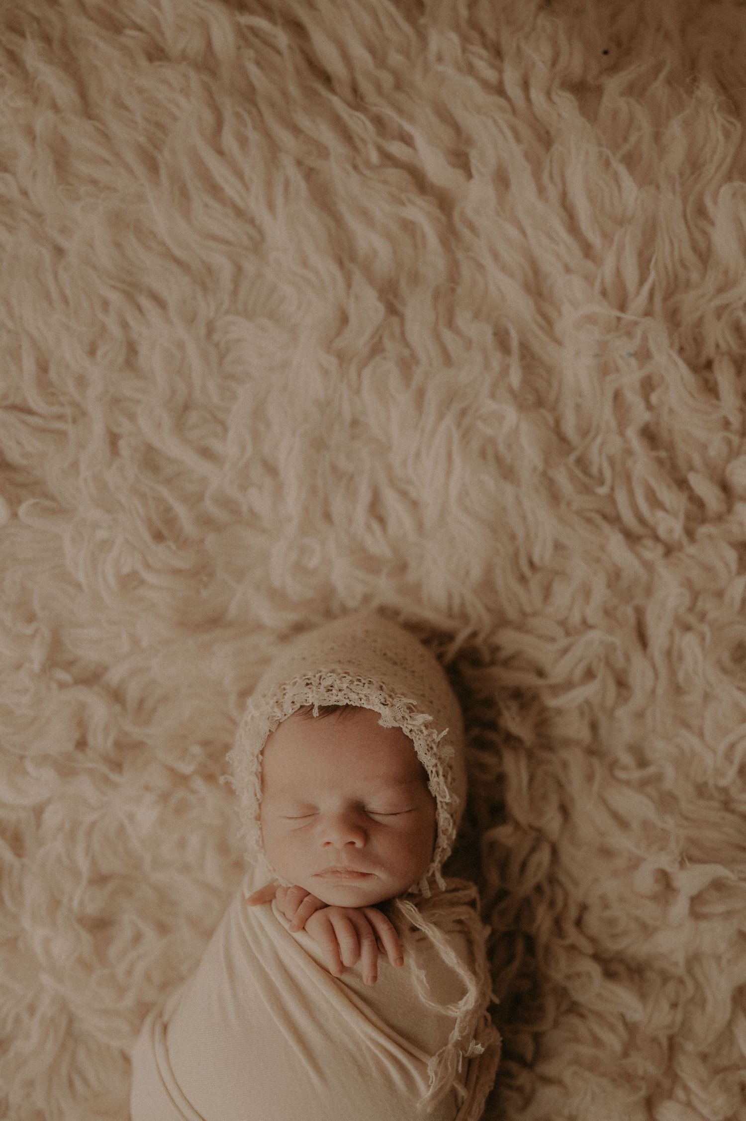 Overhead view of peaceful newborn sleeping on fluffy white rug wearing delicate bonnet.