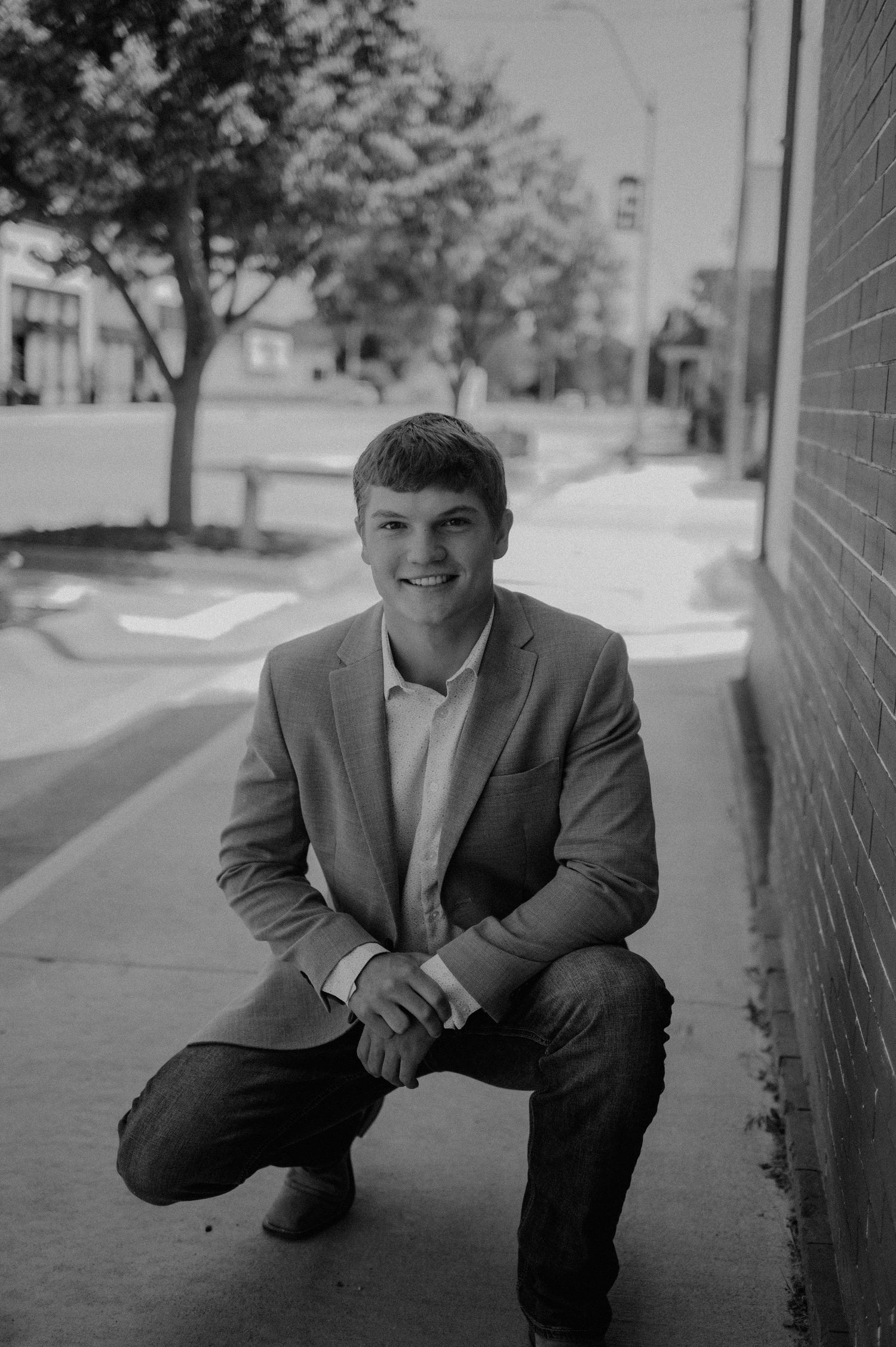 Black and white portrait of someone in a suit jacket crouching on a sidewalk with a brick wall and tree in the background.