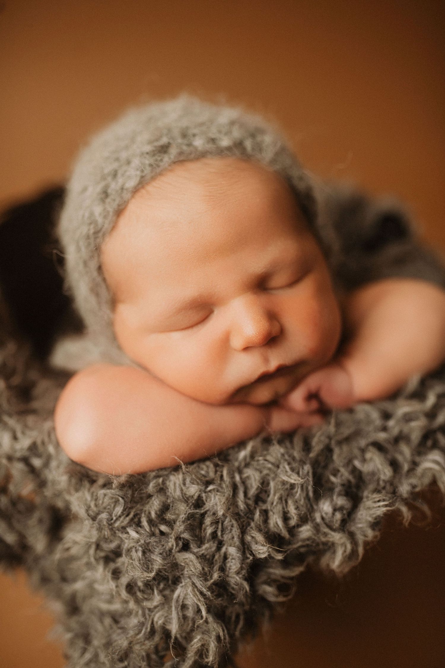 Sleeping newborn wearing gray knit bonnet resting on fluffy textured fabric.