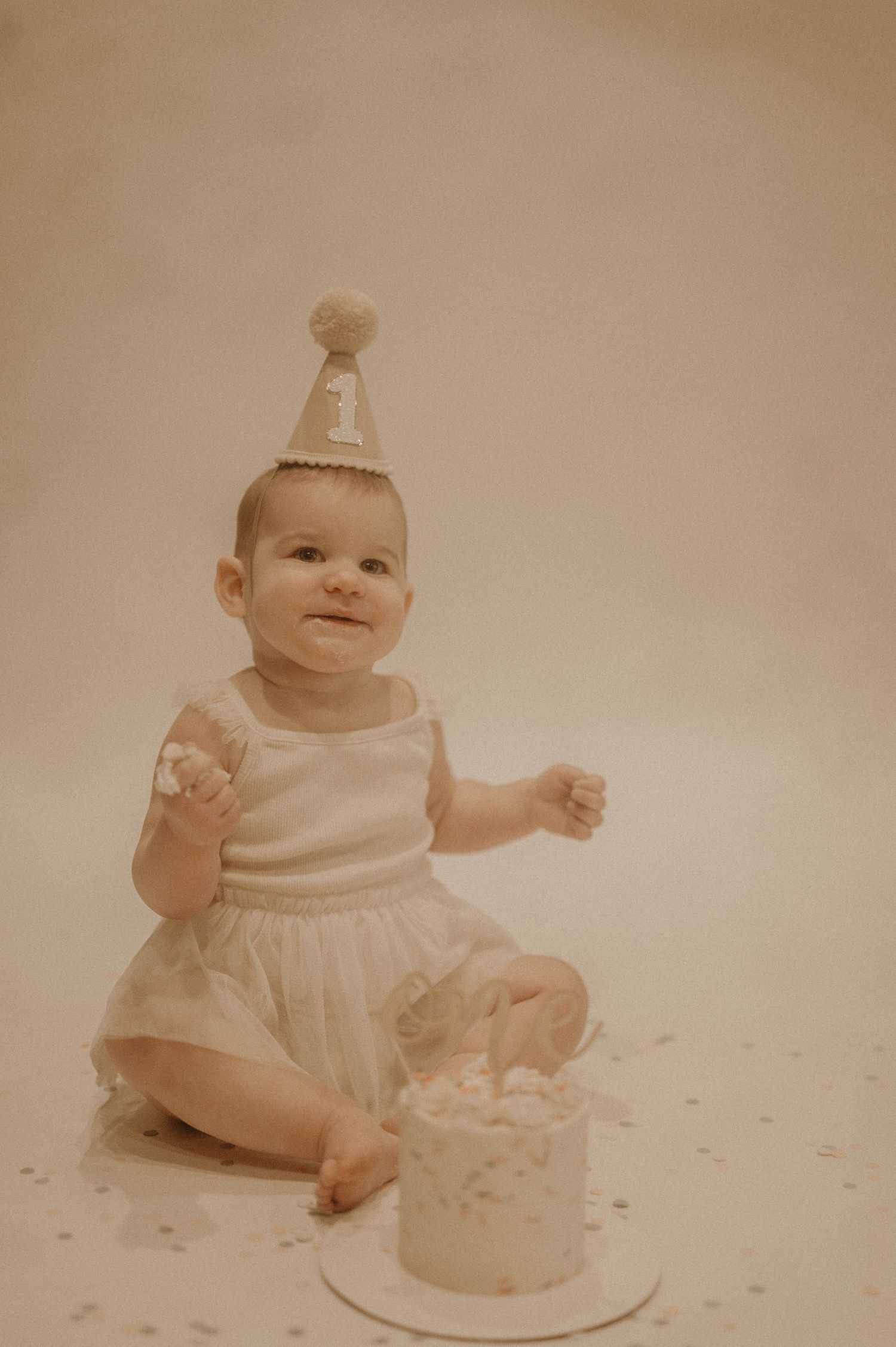 A sepia-toned photo series shows a baby in white clothing sitting with a birthday cake during a cake smash photoshoot.