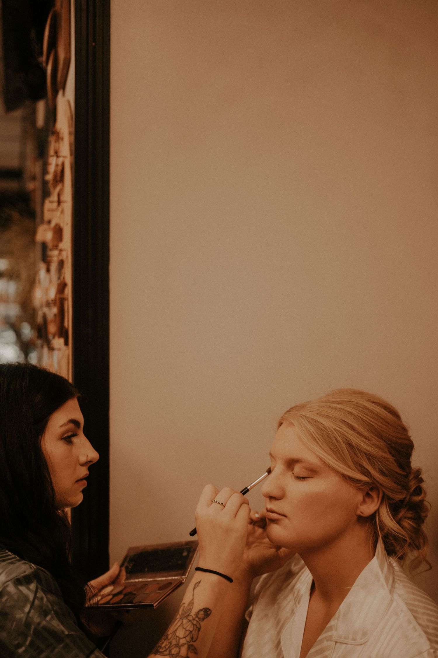A makeup artist applies eye makeup in a dimly lit room with warm lighting.