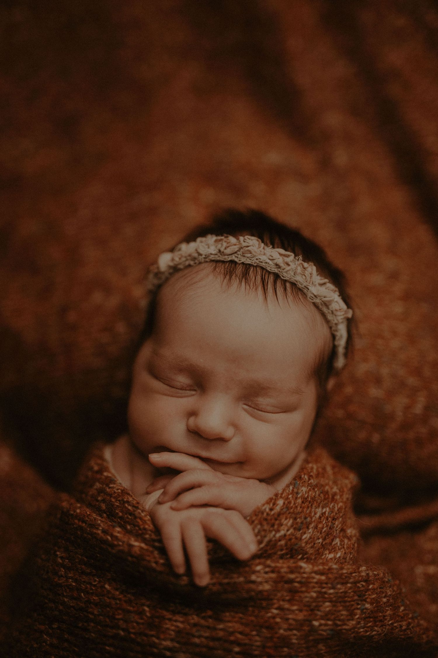 A sleeping newborn baby wrapped in a brown knitted blanket in a warm sepia-toned photograph.