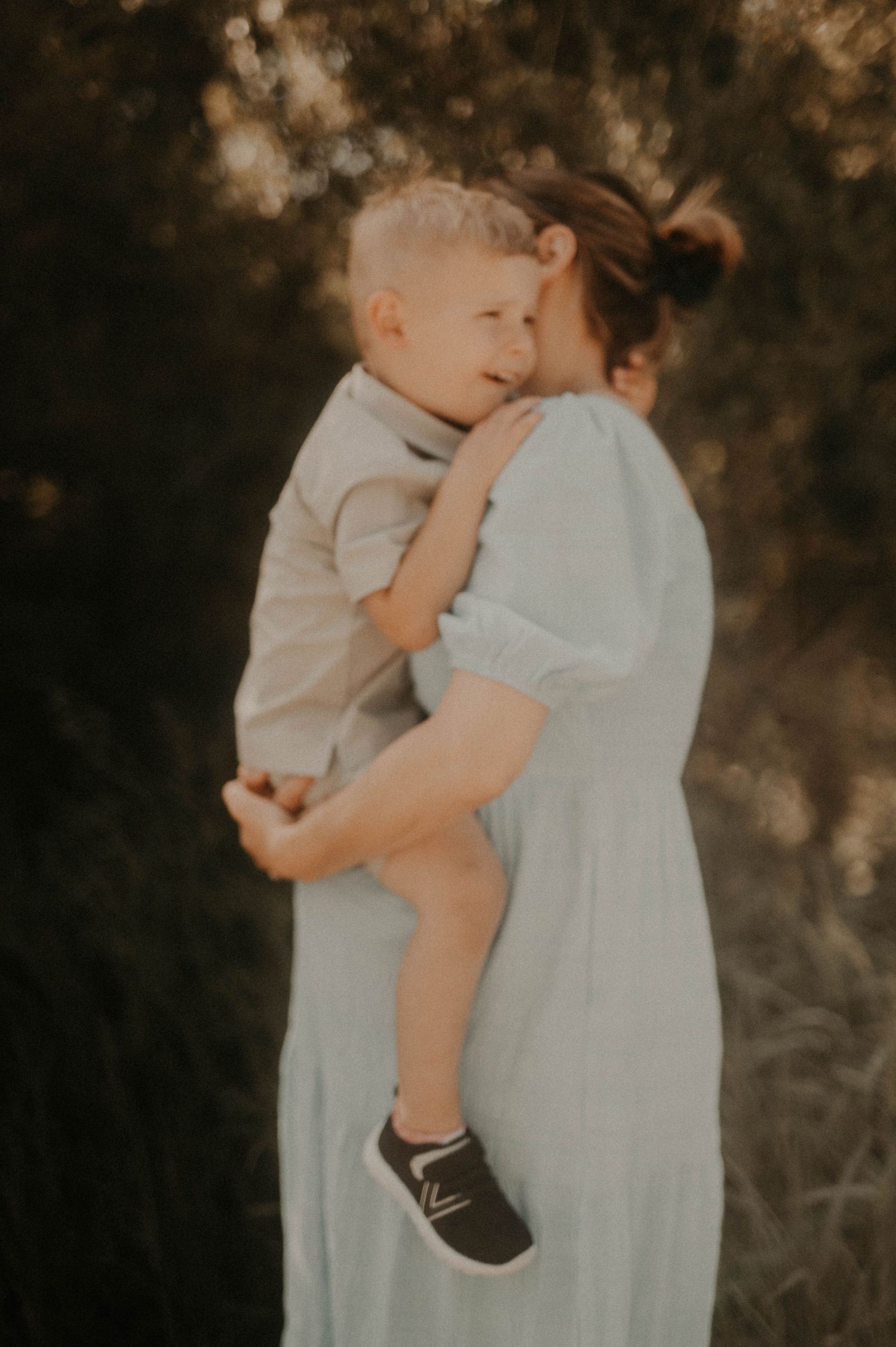 A tender outdoor portrait in soft lighting shows an intimate embrace between parent and child in a light blue dress.