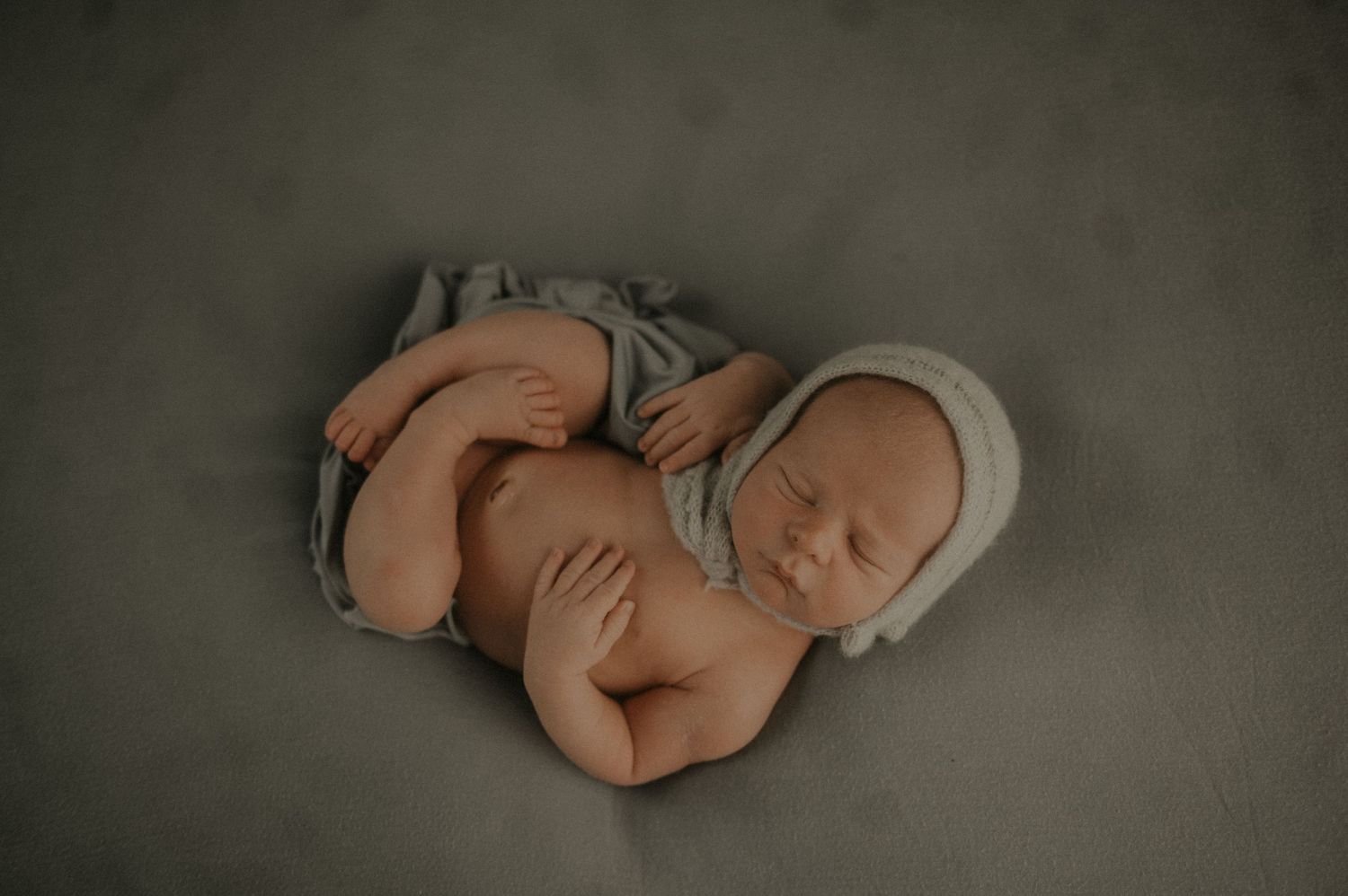 Sleeping newborn baby curled up in soft grey blanket against neutral backdrop.