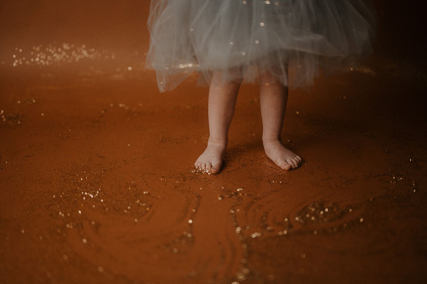Bare feet stand on a rustic orange floor beneath a flowing white tulle skirt with sparkles scattered around.