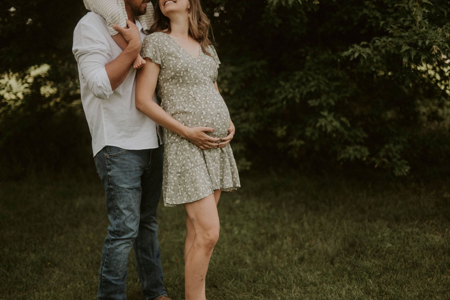 A couple embraces in a romantic pose on a grassy outdoor setting during a maternity photoshoot.