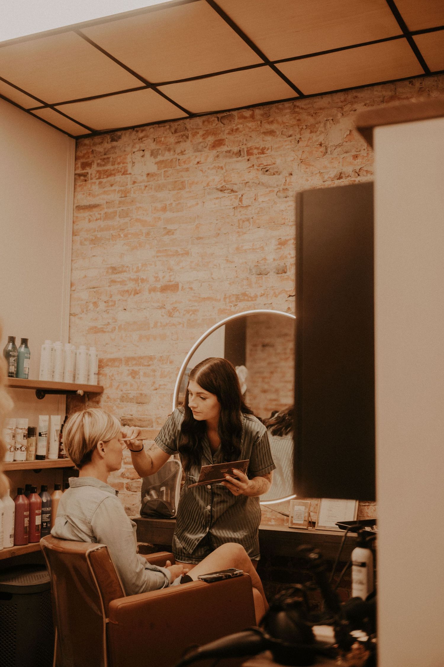 A beauty professional applies makeup in an intimate salon setting with exposed brick walls.