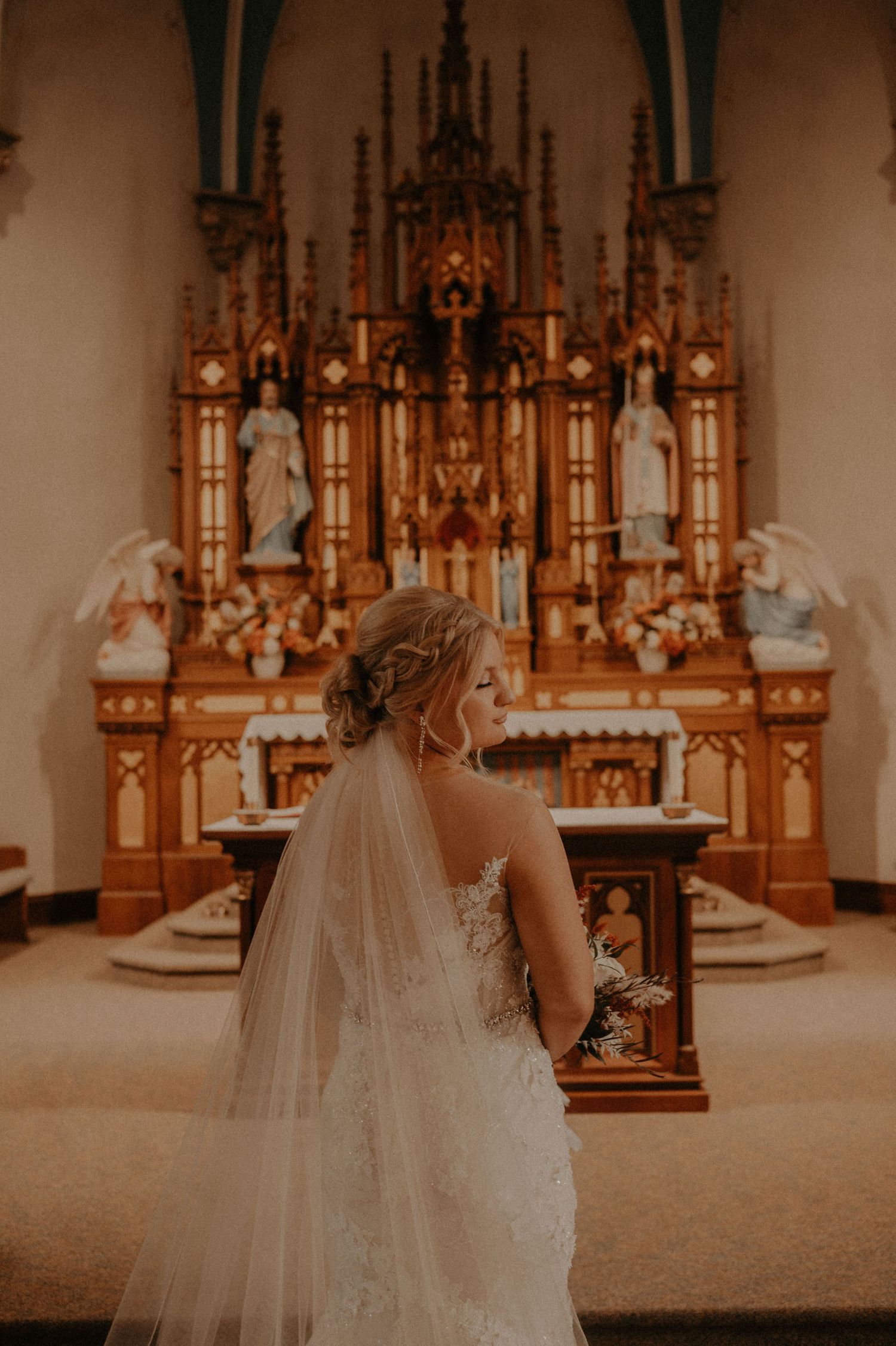 Cathedral length veil flows behind bride's elegant lace wedding gown near ornate altar.