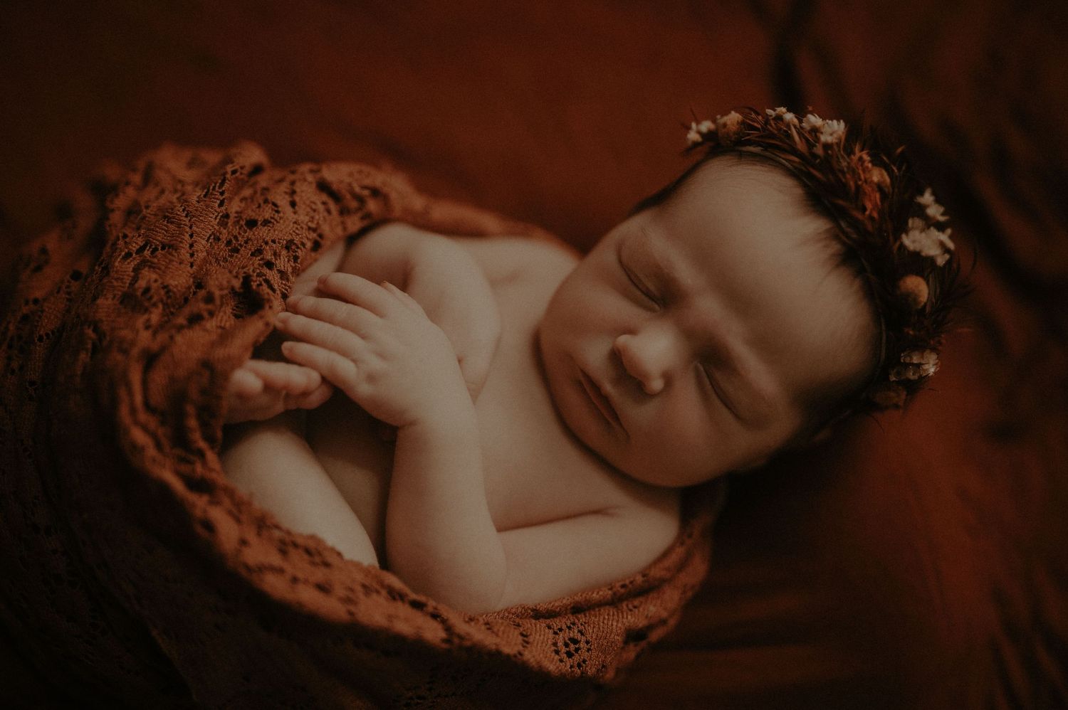 A sleeping newborn baby wrapped in orange lace fabric wearing a delicate floral crown in warm moody lighting.